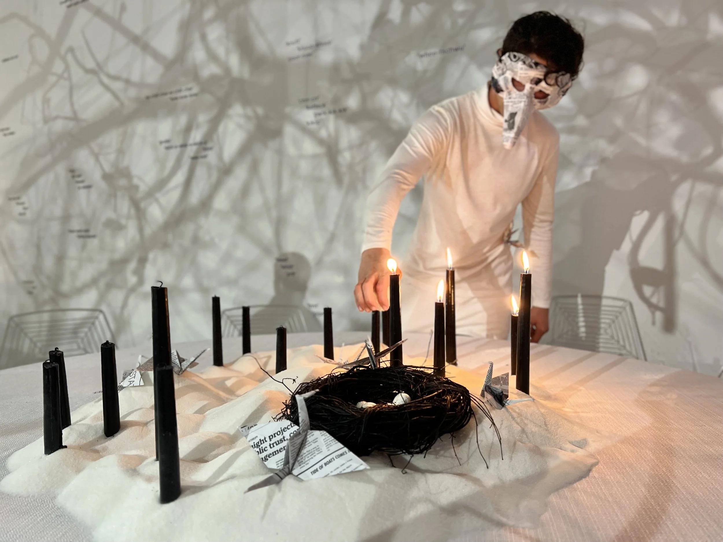 A person wearing a newspaper mask stands behind a table with lit candles arranged on it. The table is covered with newspaper and clouds of shadows and text are cast on the wall behind them.