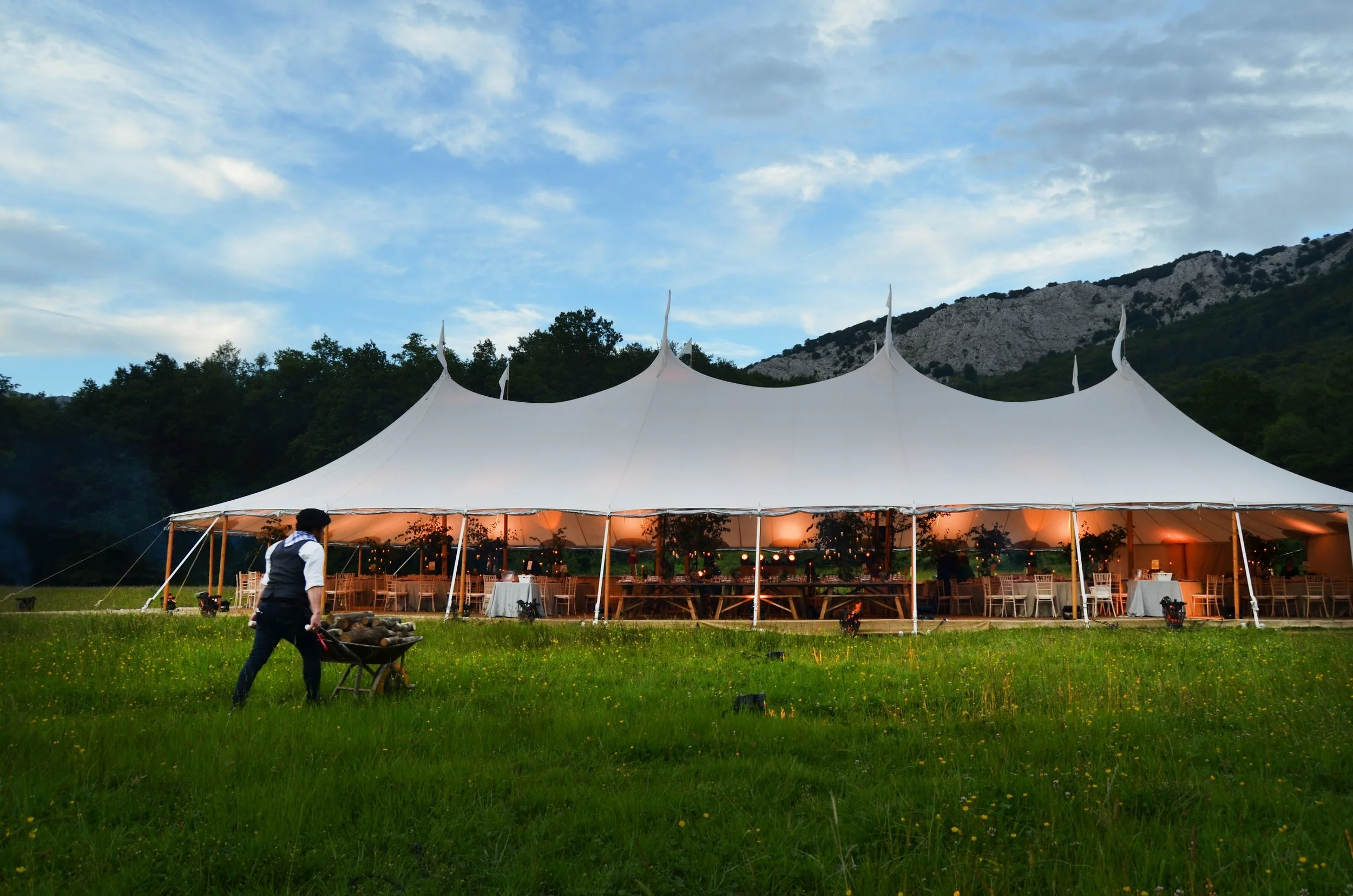 An outdoor event setup under a large white tent with string lights, surrounded by greenery and mountains in the background during evening.