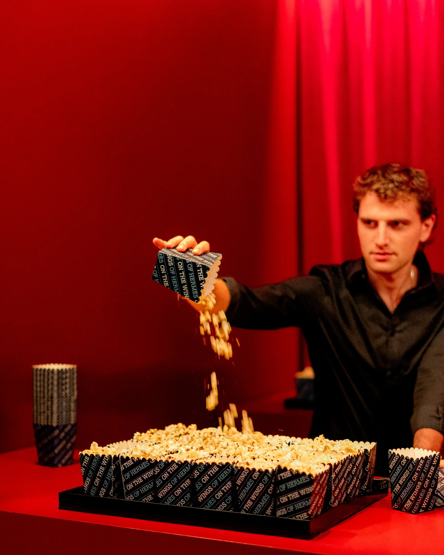 A person with curly hair holding a tray with falling popcorn against a red background.