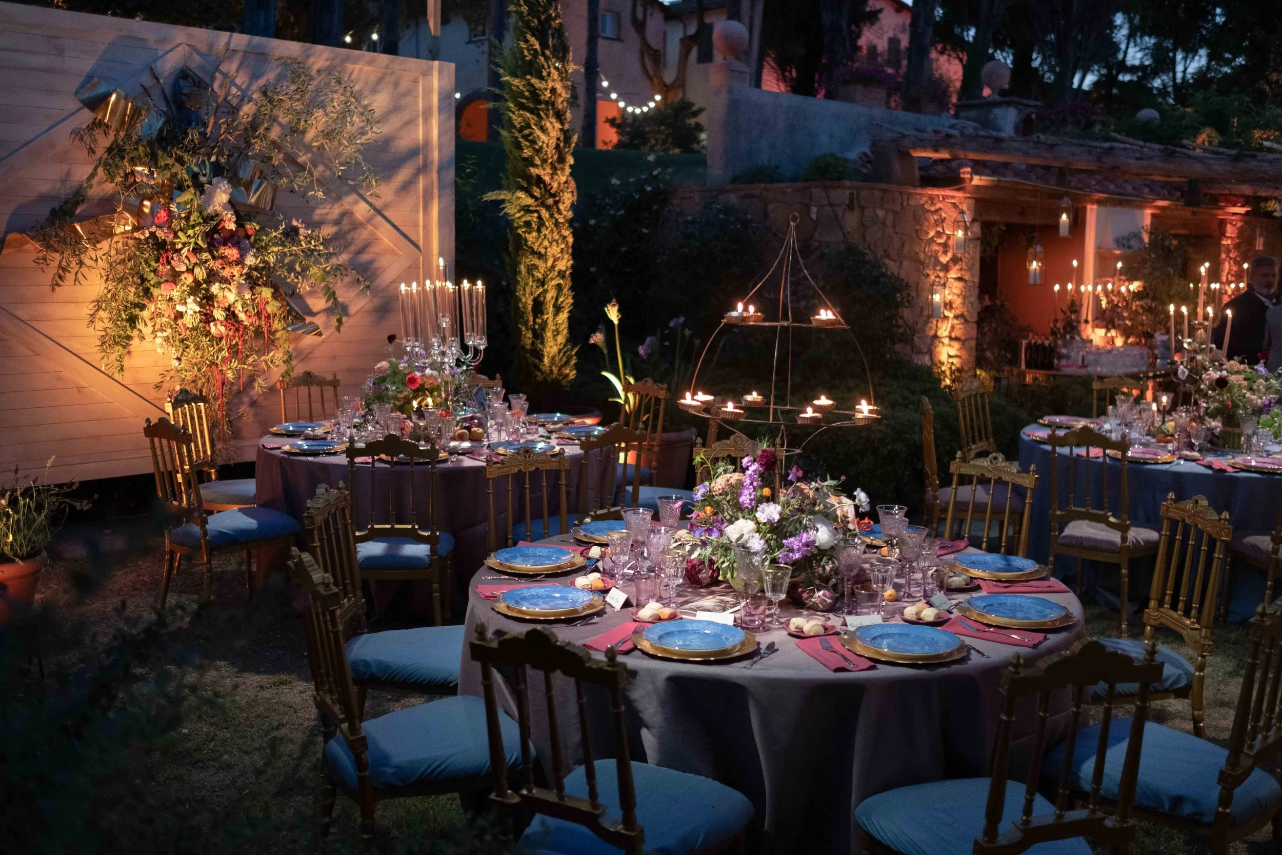 Outdoor evening wedding reception setup with decorated tables, floral arrangements, candles, and string lights in a garden setting.