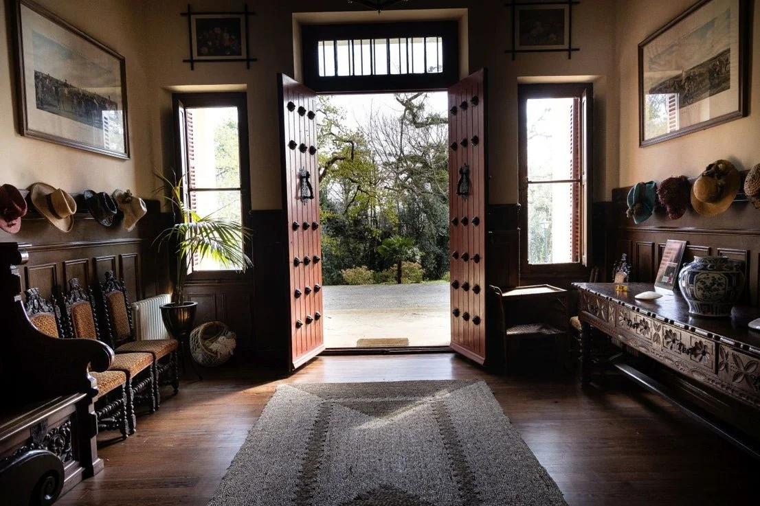 Interior view of the Palacio de Arbaisenea with a wooden-floored entryway with open double doors leading outside to trees, with hats hanging on the wall and furniture including chairs, a side table, and a decorative cabinet.