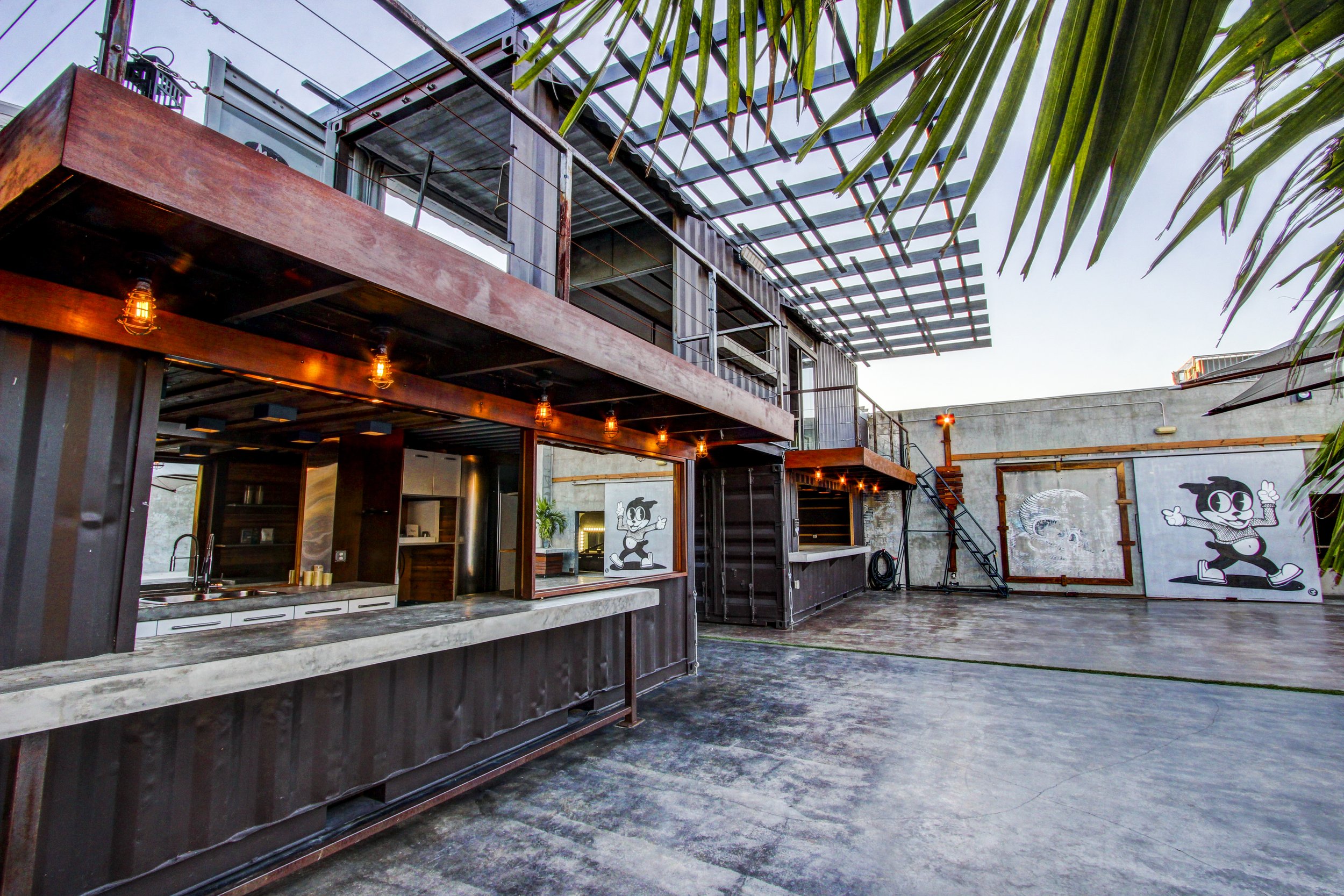 Outdoor patio area with a wooden bar, string lights, a mural of Mickey Mouse, and palm tree leaves overhead.