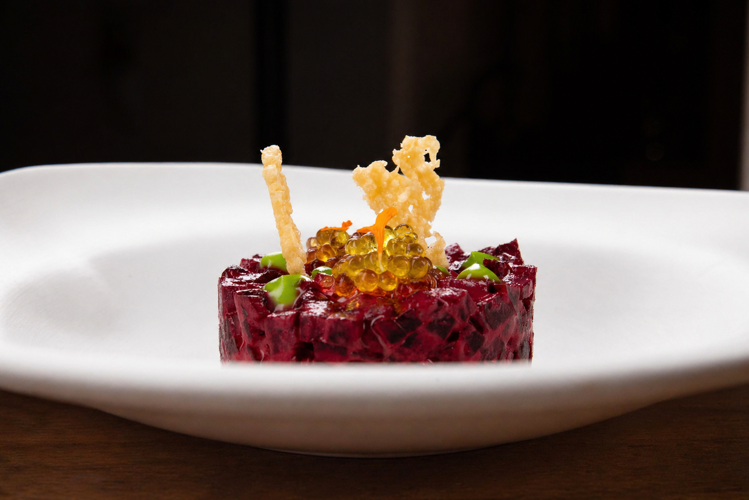 A gourmet dish with a cube of red beet tartare, topped with orange fish roe, green accents, and crispy garnishes in a white bowl on a wooden surface.