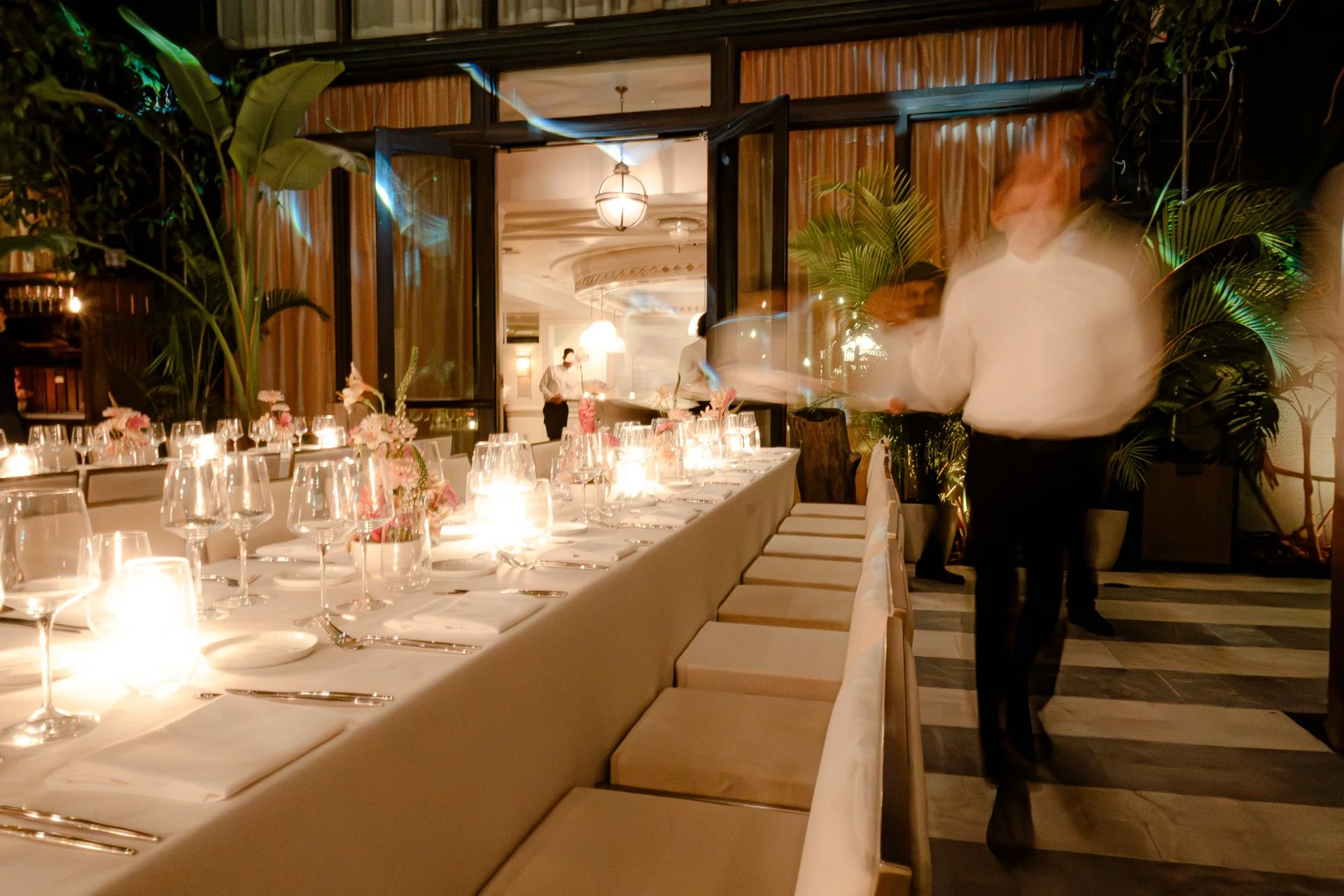 A dimly lit restaurant with a long table set with fine dining ware, candles, and floral centerpieces. Large green plants and windows with curtains are in the background. There are a few people in the background, with one person in white attire and others partially visible, moving around.