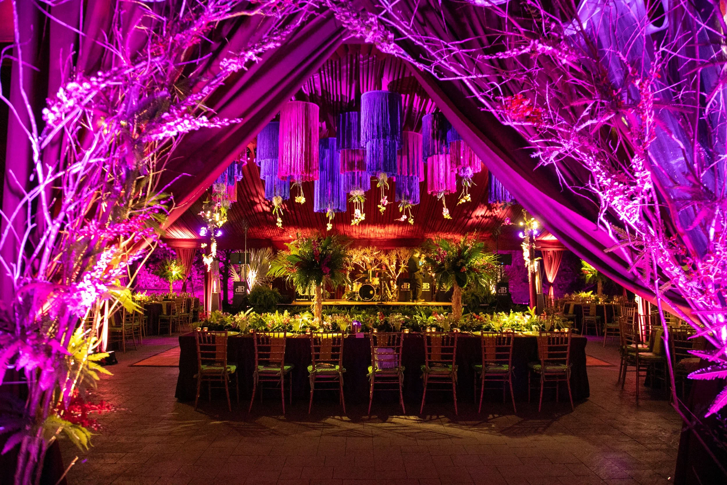 Decorative event space illuminated with purple and pink lighting, featuring hanging fabric or tassels, floral arrangements, and tables with chairs.