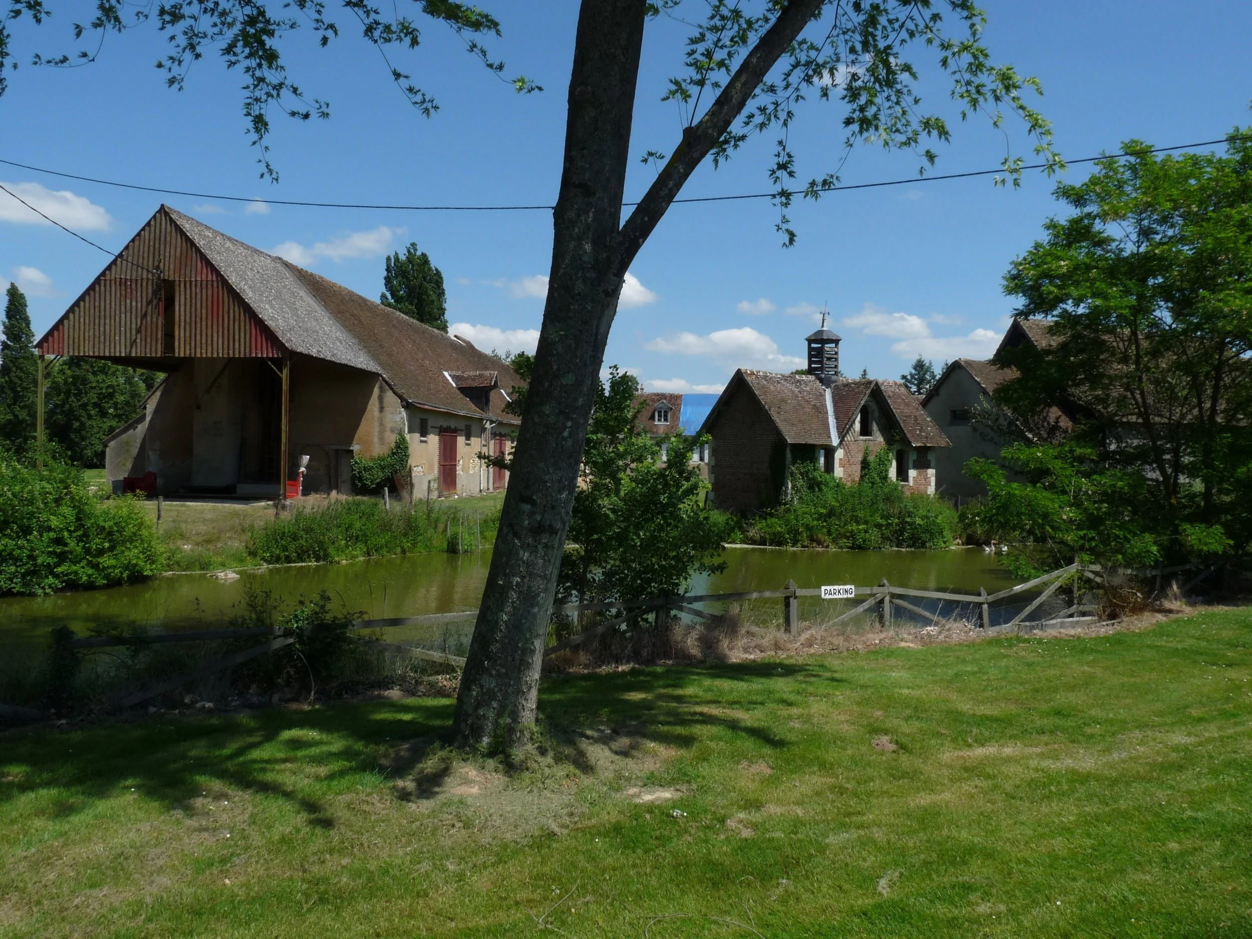 A scenic view of a small group of farm buildings that can house special events and located  by a river, with trees, a barn, houses, and a church steeple under a bright blue sky with clouds.