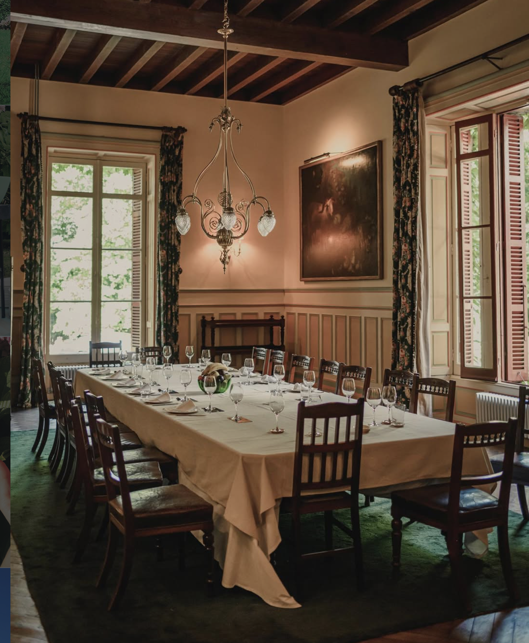 A dining room at the Palacio de Arbaisenea, with a long table set for a meal, featuring white tablecloth, wine glasses, and a fruit bowl in the center. The room has large windows with floral curtains and wooden shutters.