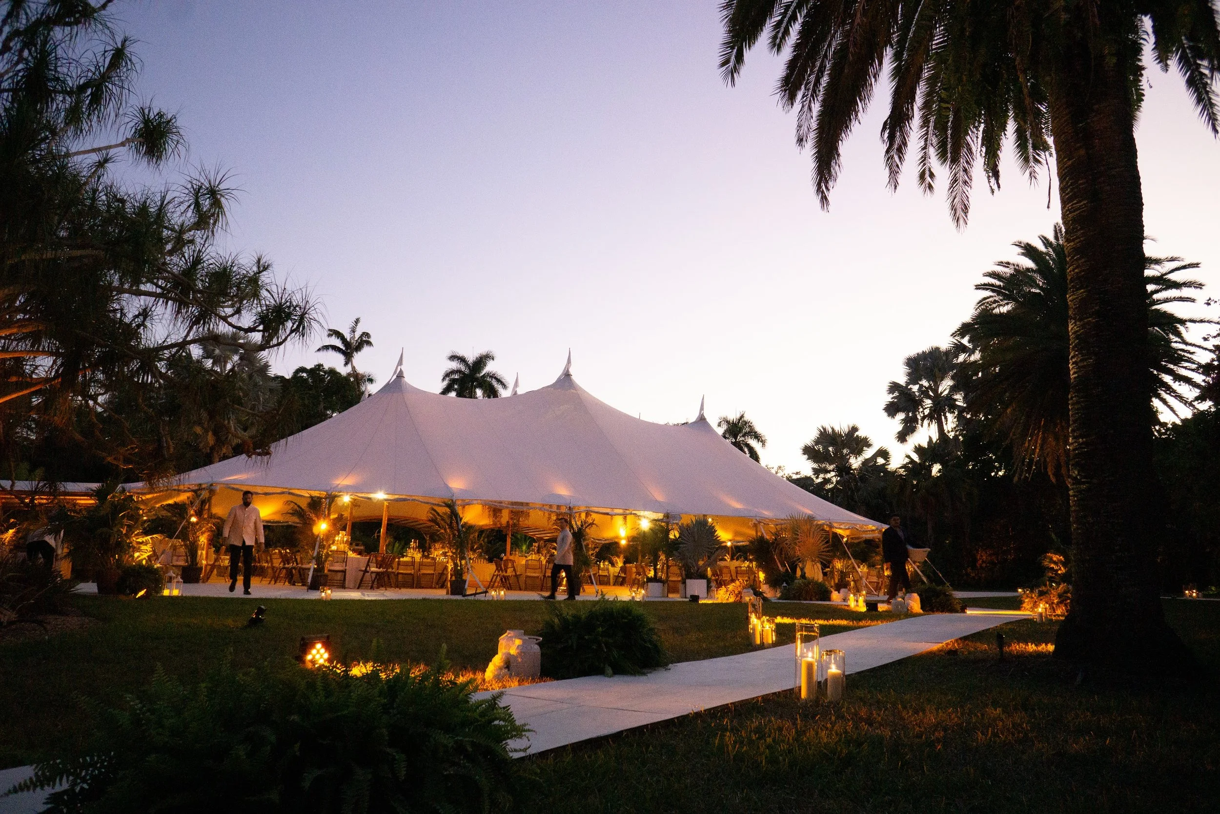 Outdoor event at dusk with a large white tent, palm trees, and string lights, with people standing and walking nearby.