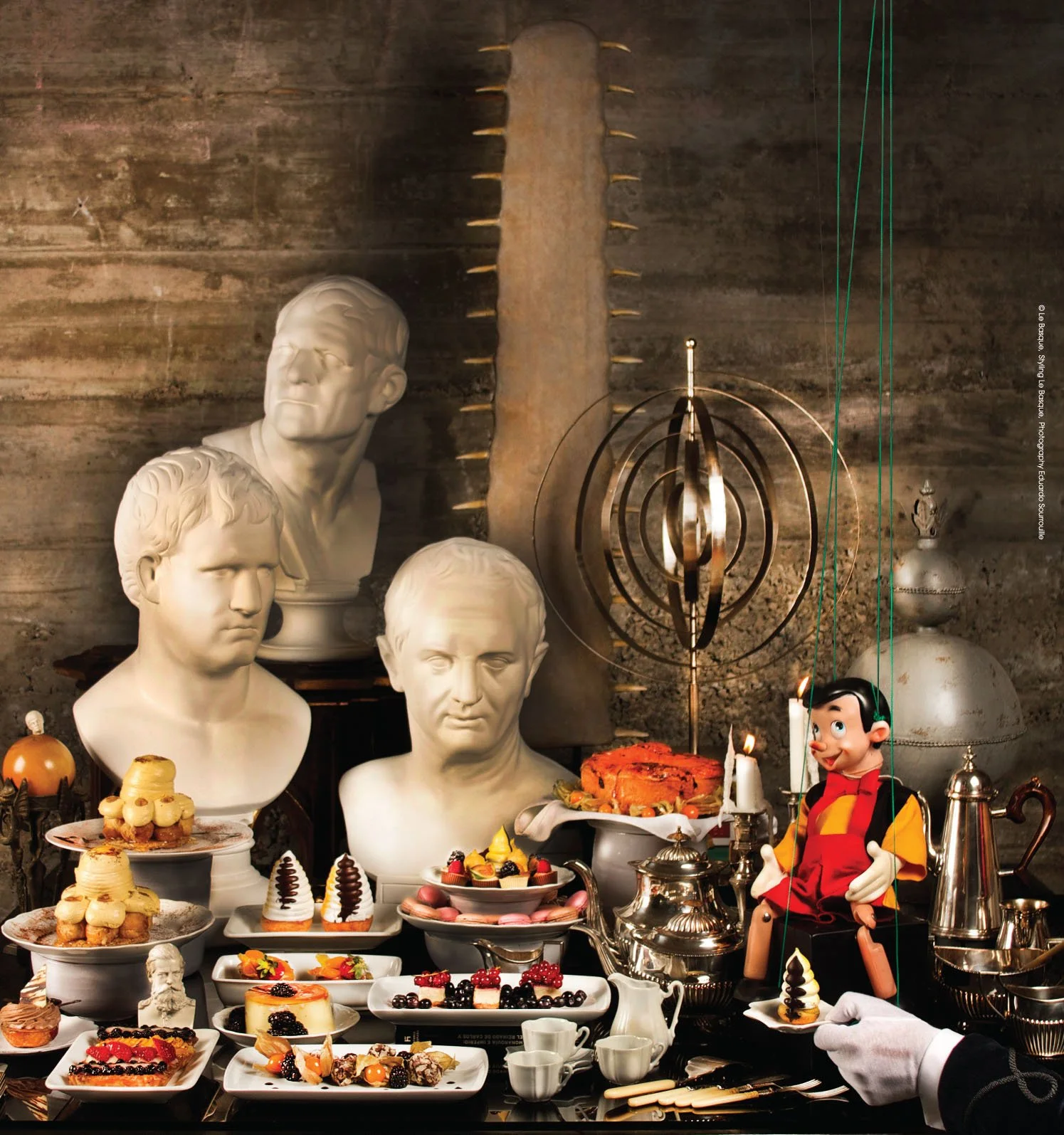A festive table with various desserts including cakes, tarts, and pastries, surrounded by bust sculptures and a Pinocchio puppet, with a dark background and vintage decor.