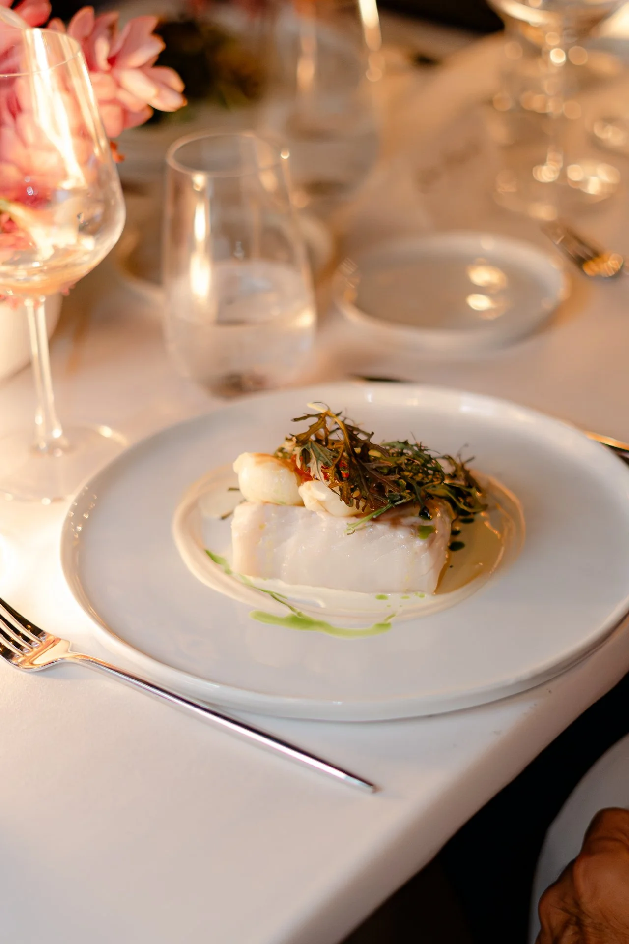 A plated appetizer with a slice of fish topped with greens on a white table. Glassware and a floral centerpiece are visible in the background.