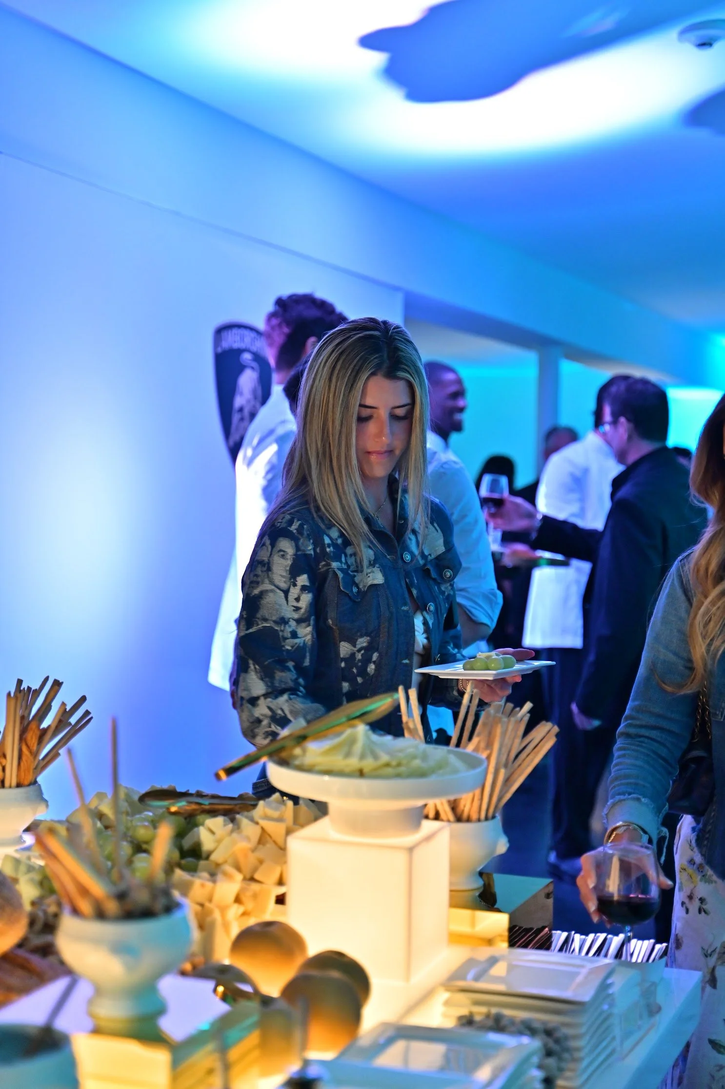 A woman in a denim jacket holding a plate of green grapes at a buffet table with cheese, crackers, skewers, and other snacks, at an indoor event with blue lighting and people socializing in the background.