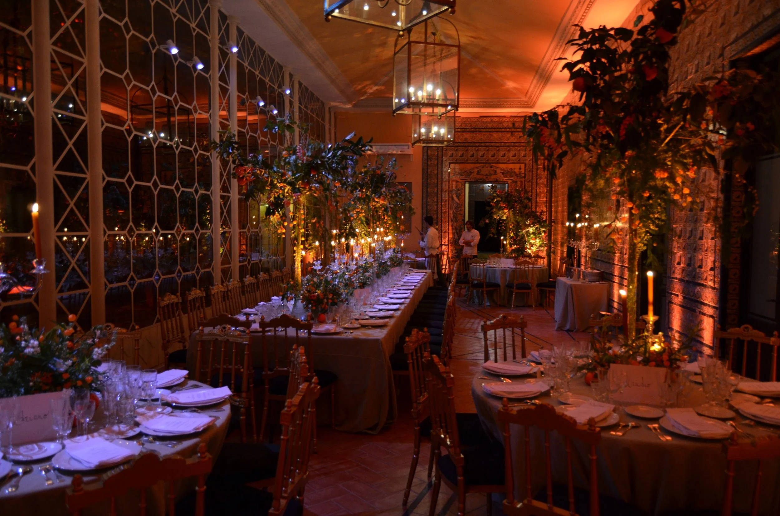 An elegant banquet hall decorated for a dinner event with long tables, floral arrangements, and warm lighting. Two waitstaff members are preparing near the table, and the room features patterned walls, chandeliers, and a decorative partition.