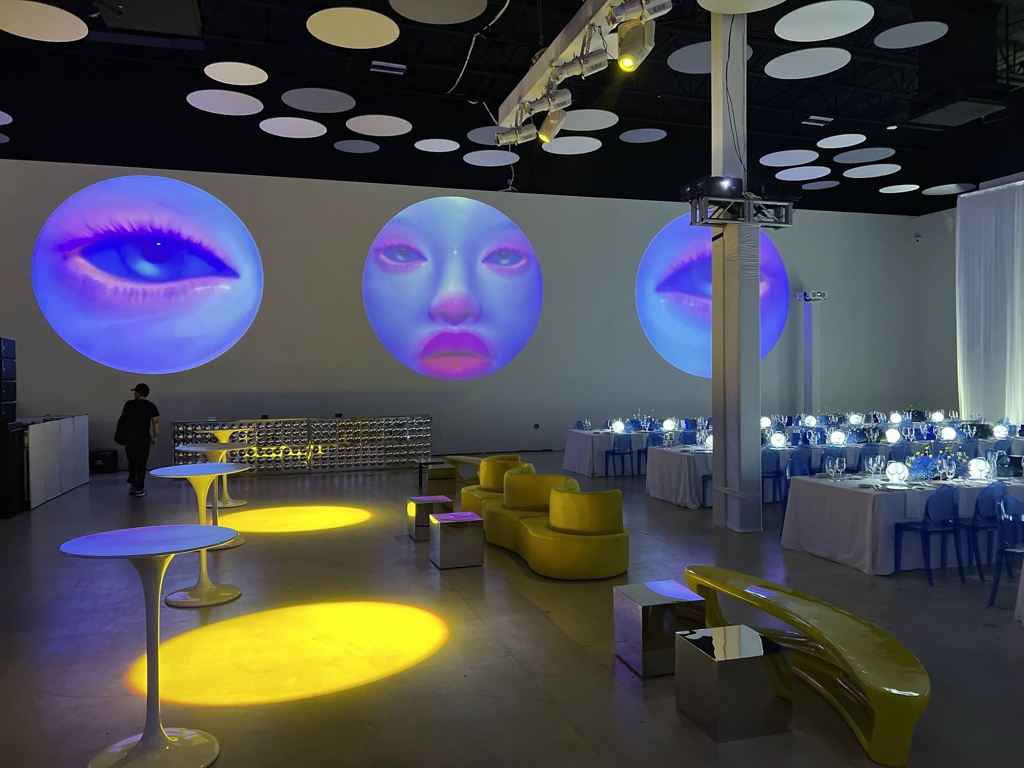 An indoor event space with large circular digital screens displaying artistic images of eyes and lips. The space features yellow and blue seating, round tables with tableware, and overhead lighting. The setting appears to be a modern, stylish venue for a gathering or celebration.