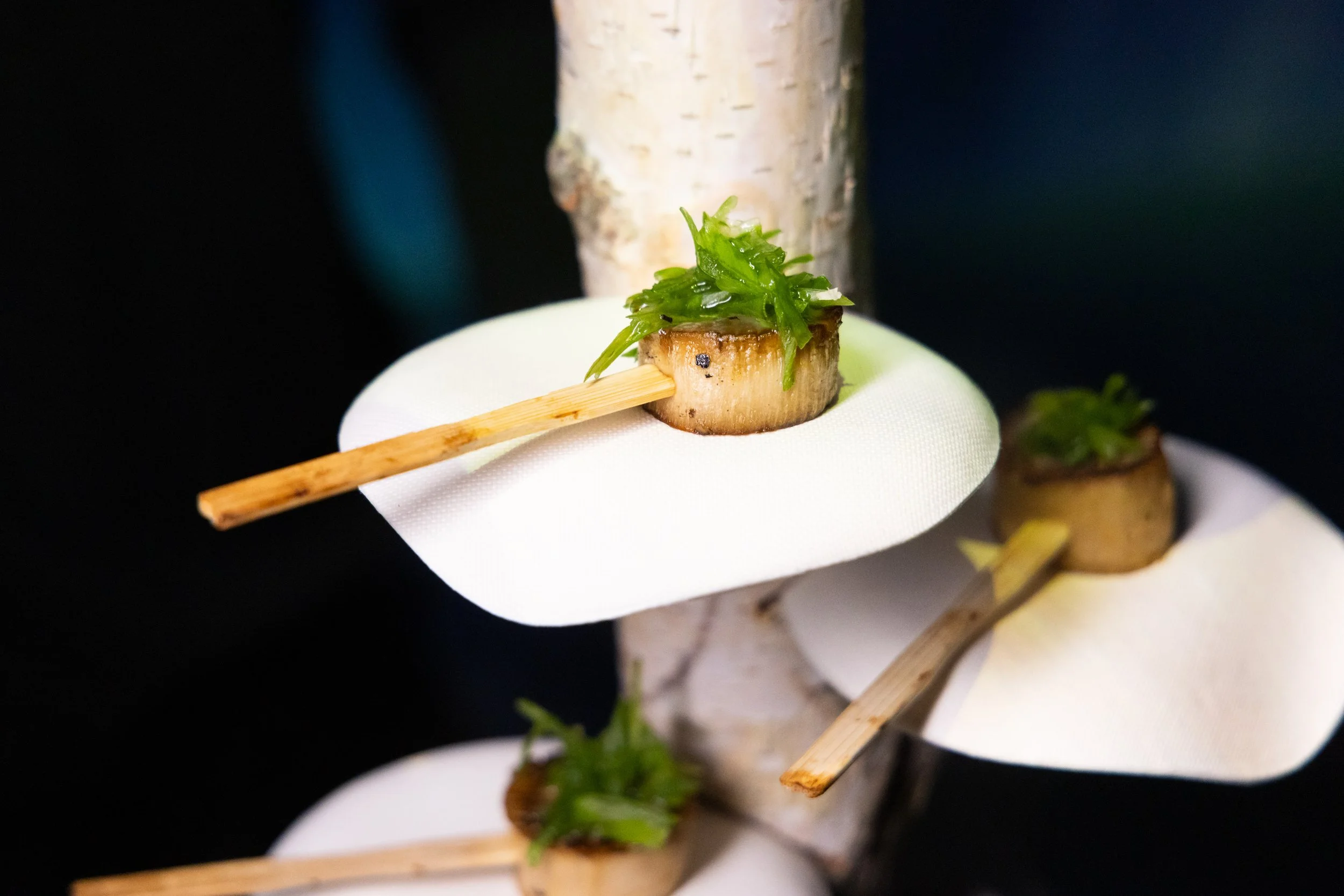 Close-up of a piece of food with green garnish held by a person wearing a white glove, with blurred background.