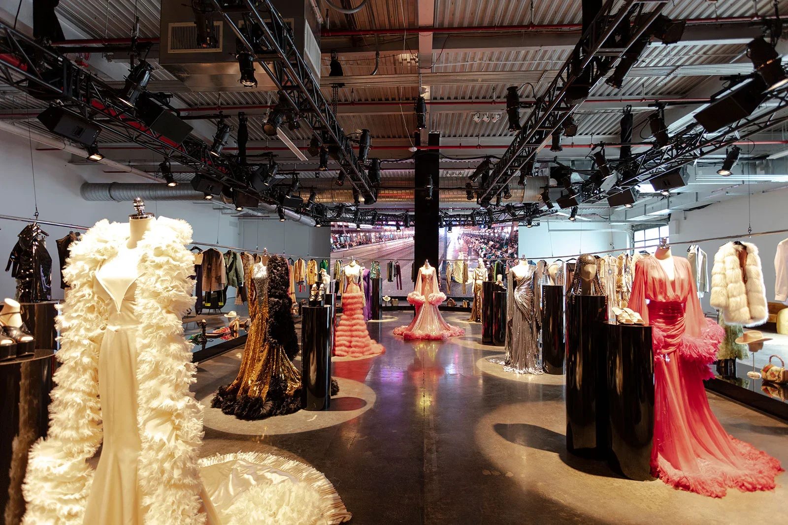 An exhibition of evening gowns and fashion clothing displayed on mannequins and hangers in a spacious showroom with black pillars and a large digital screen in the background.
