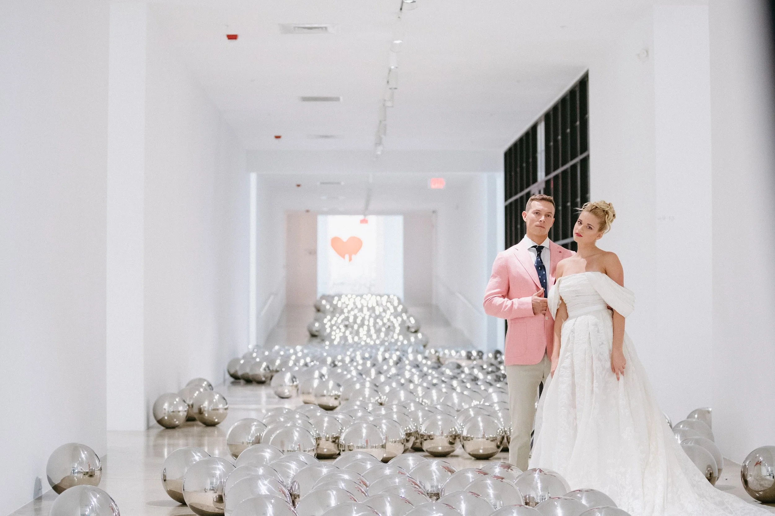 A man and woman in wedding attire standing together in a modern, white hallway decorated with shiny silver balloons and a string of lights. The man is dressed in a pink suit with a white shirt, navy tie, and pocket square. The woman is wearing a strapless white wedding gown and has her hair styled in an updo with a headpiece. A large heart symbol is visible on the wall in the background.