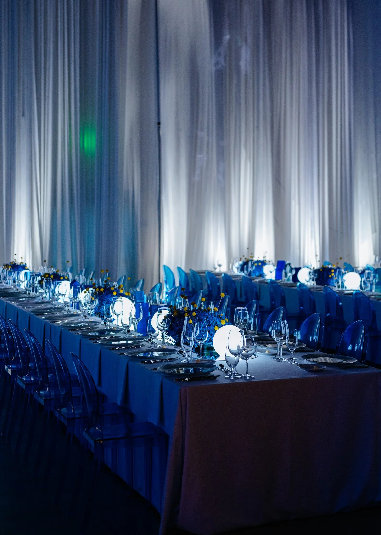 A long banquet table set for a formal event with white tablecloths, glassware, and floral centerpieces illuminated with blue lighting, with a backdrop of tall white curtains and soft ambient lighting.