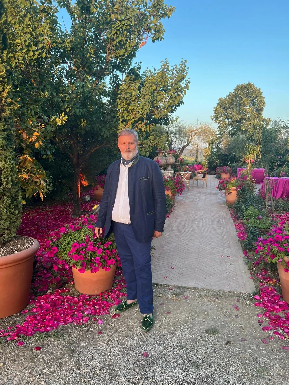 A man standing on a paved garden path surrounded by colorful potted flowers and lush green trees, with outdoor furniture and a clear blue sky in the background.