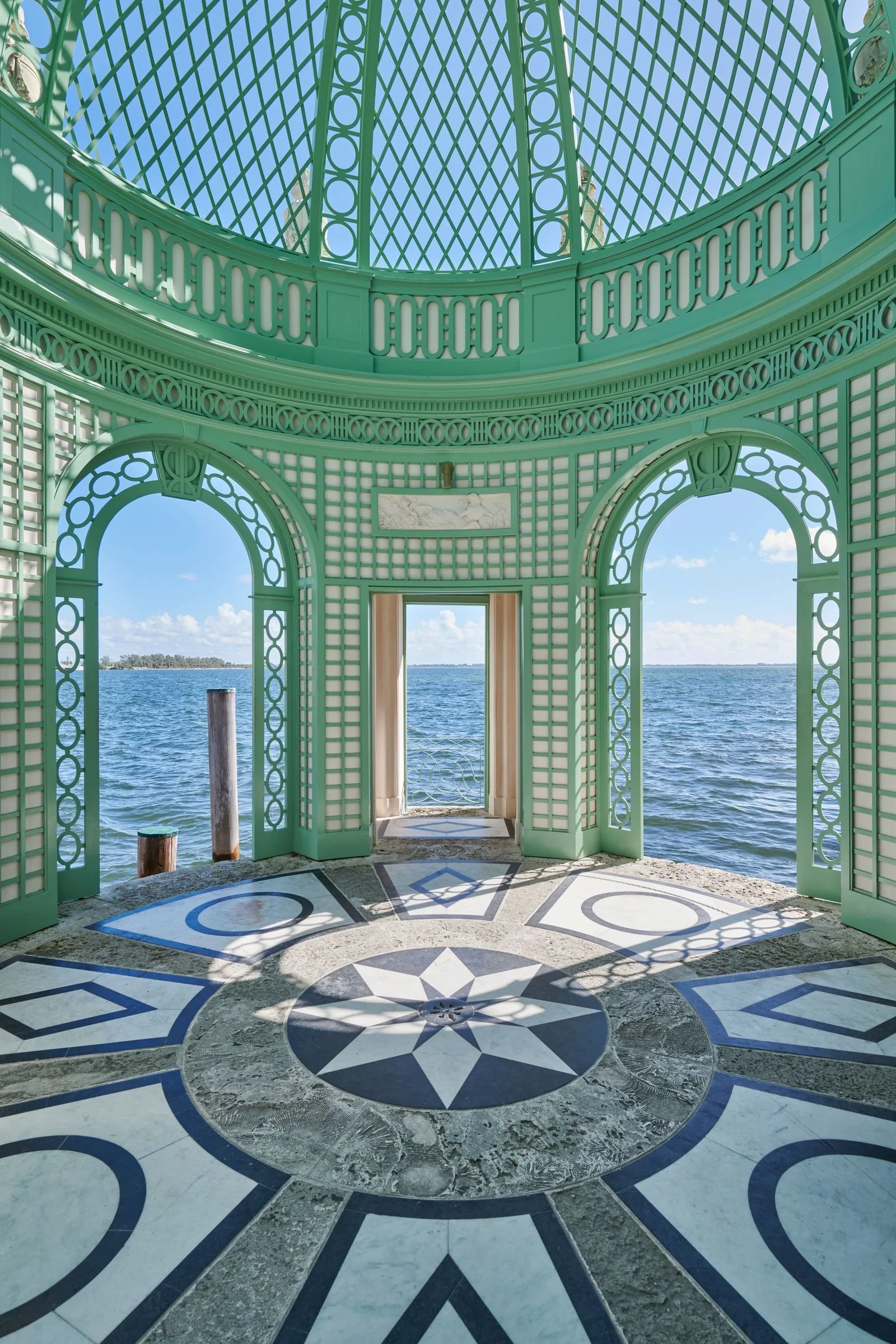 View of a seaside pavilion with teal-colored decorative openwork walls and ceiling, overlooking the water and sky, featuring a patterned floor with dark and light contrasting designs.