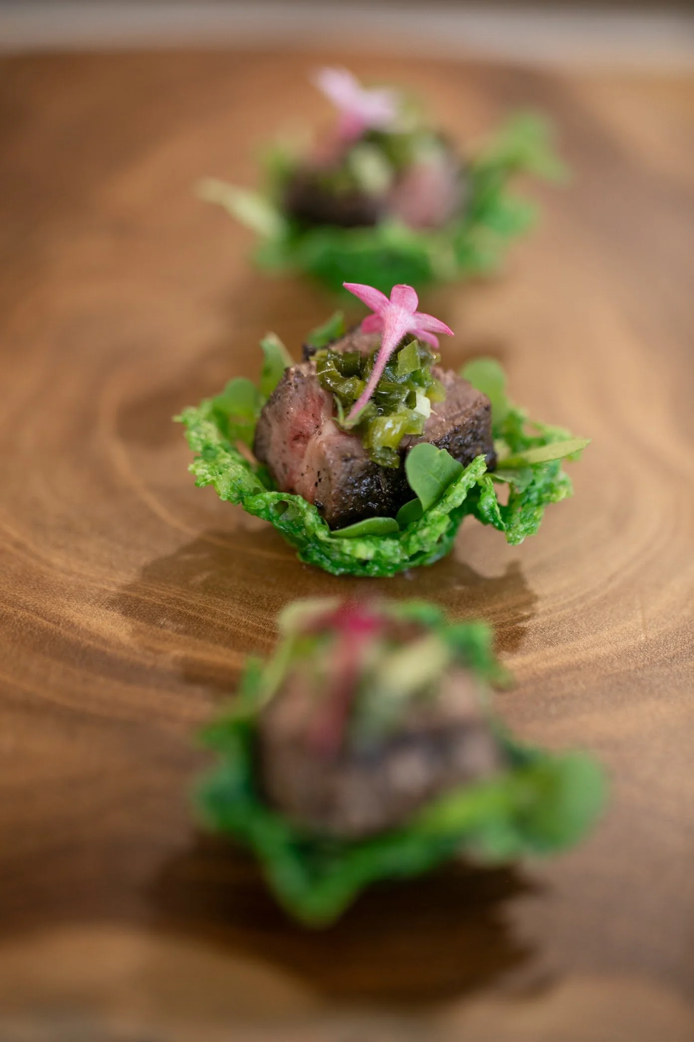 Microgreens with a pink flower on top, served on a small mound of green leaves, arranged on a wooden surface.