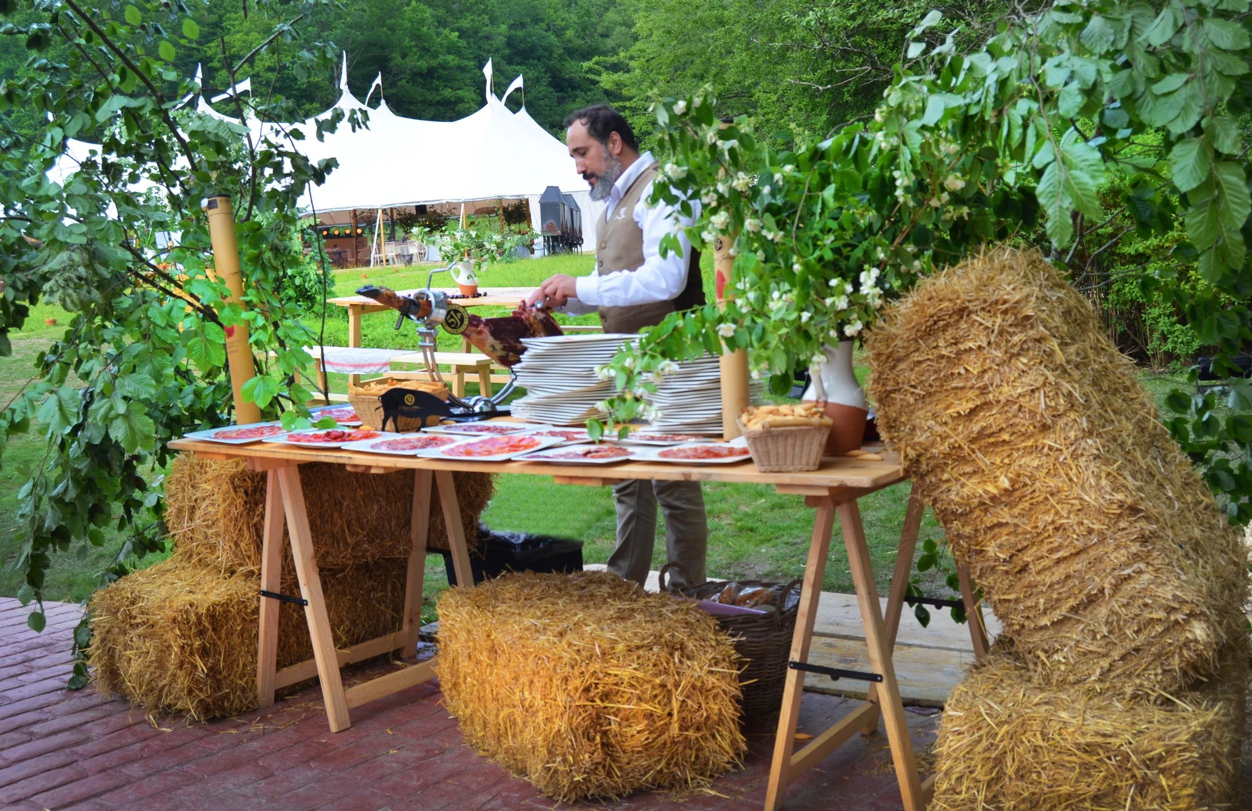 A man preparing food outdoors at a farm-themed event with hay bales, trees, a large white tent, and a grassy area in the background.