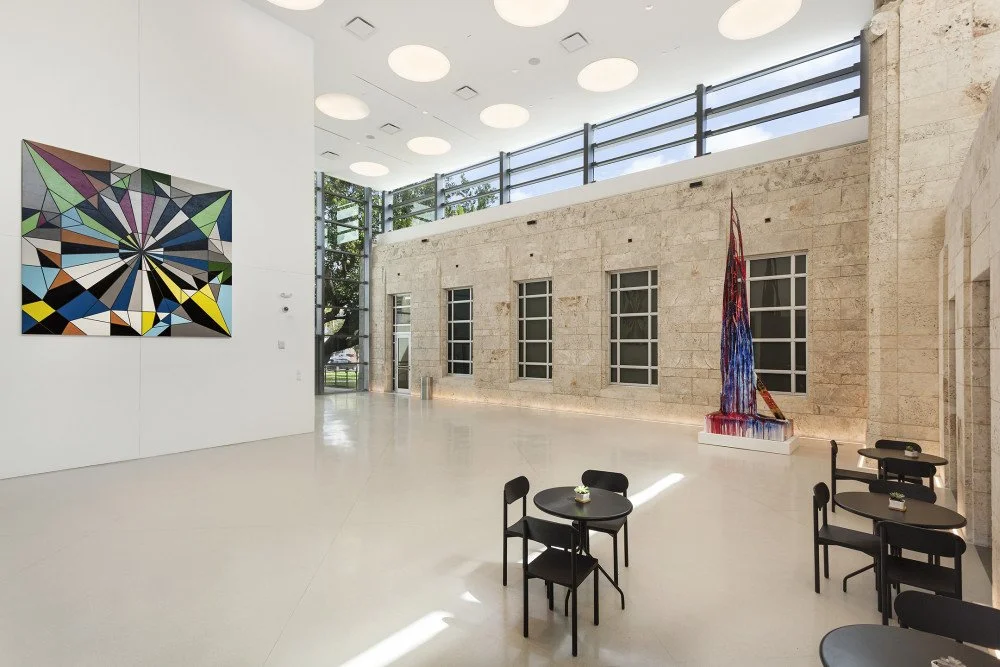 Modern art gallery with geometric paintings and sculpture, small tables with chairs, large windows, and a bright, spacious interior.