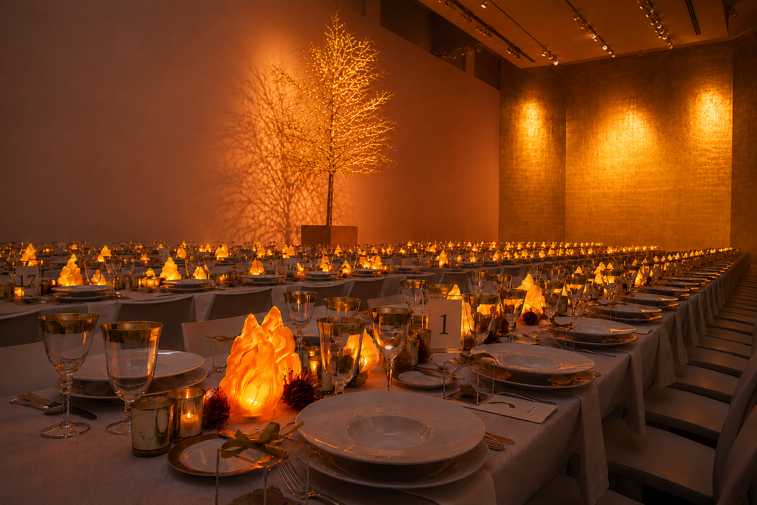 A museum gallery filled with long tables decorated with glowing orange flame candelabras, a  golden tree, and a spectacular gold leaf wall.