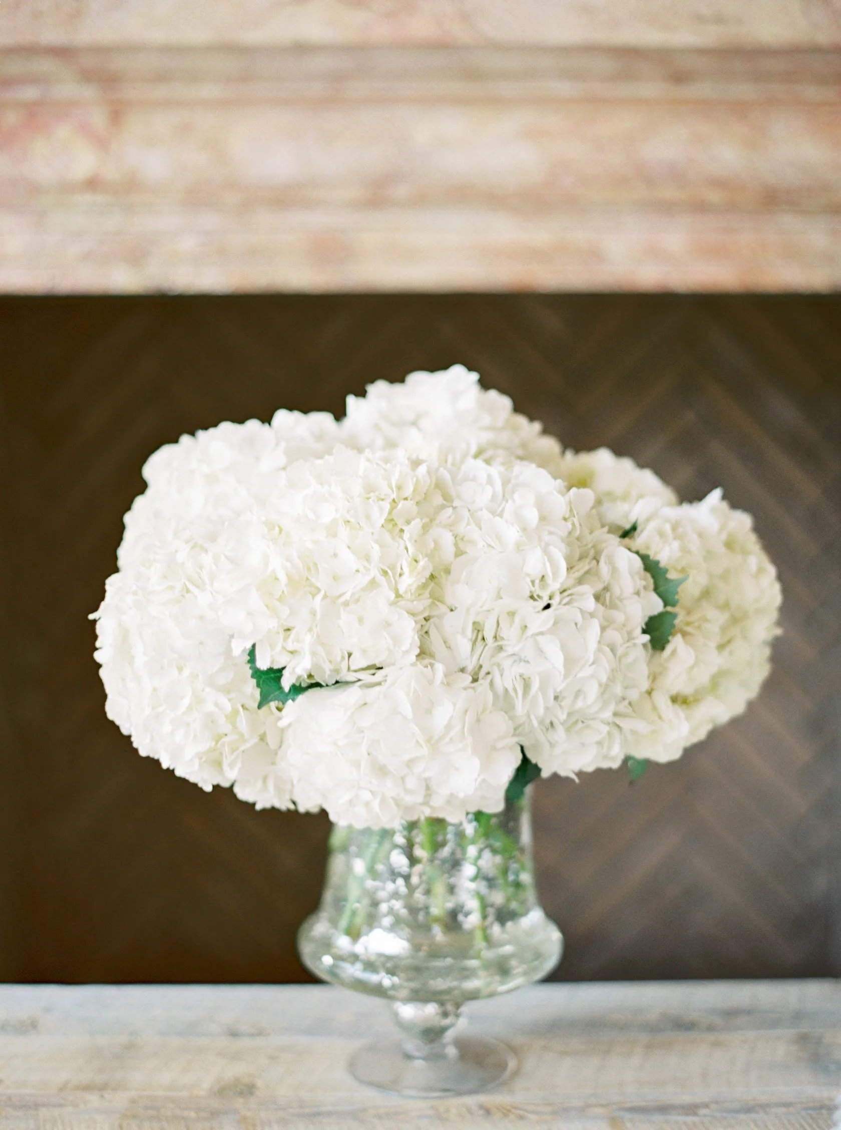 A bouquet of white hydrangea flowers arranged in a tall, elegant glass vase on a wooden surface.