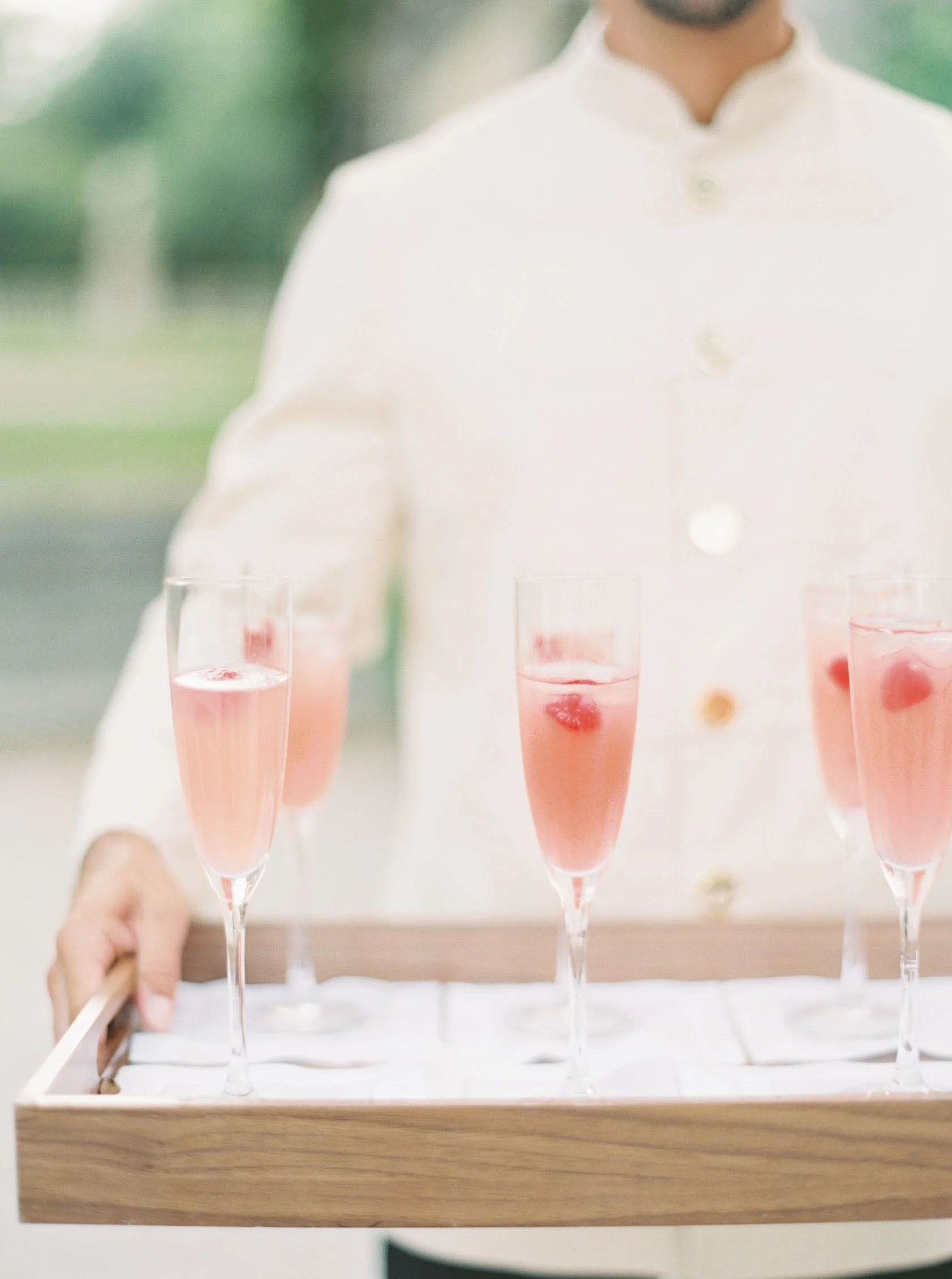 Person in white chef jacket holding tray with five glasses of pink beverage garnished with raspberries.