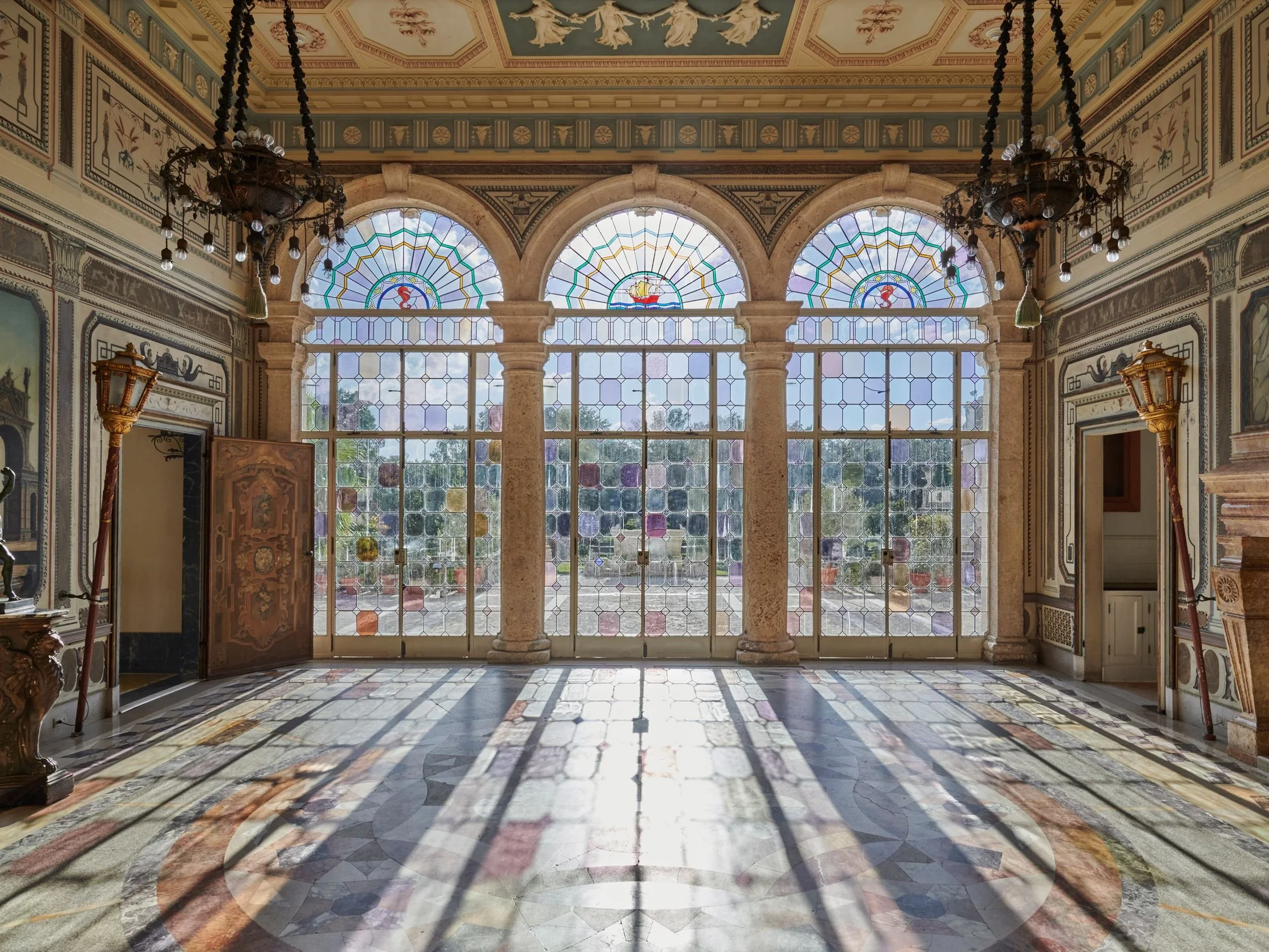 An ornately decorated interior room with large stained glass windows and intricate ceiling and wall designs, sunlight casting colorful shadows, and antique fixtures.