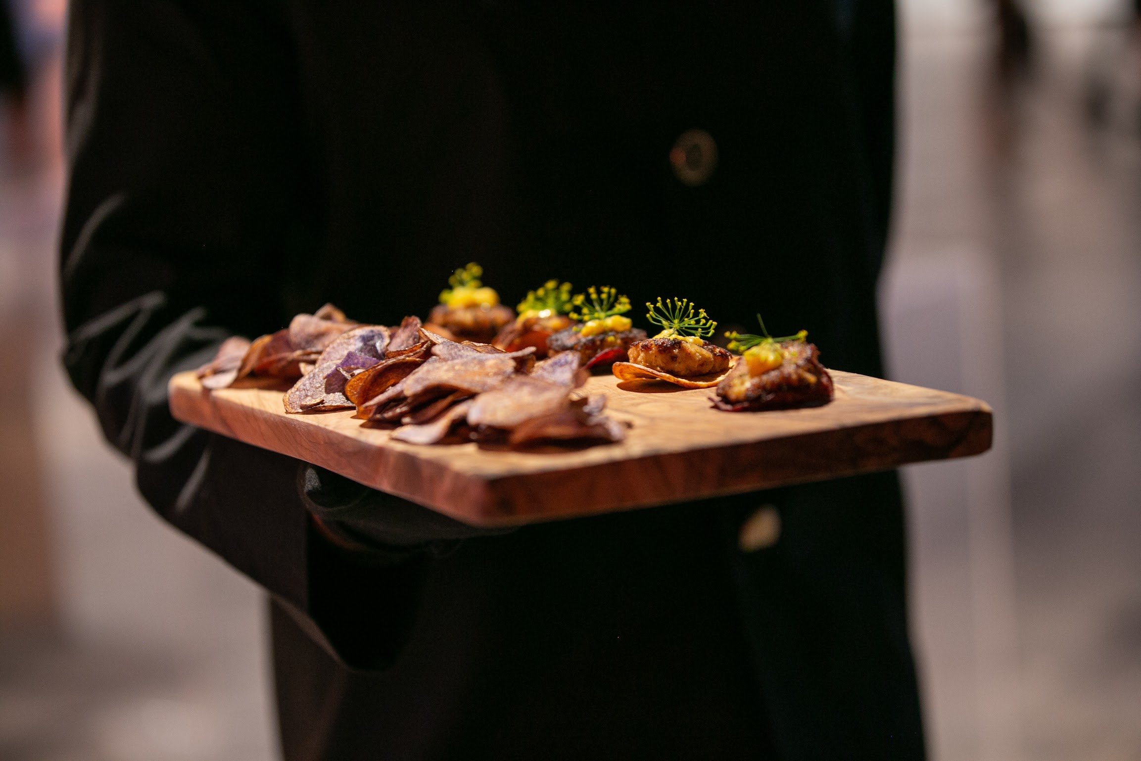 A person in a black suit holding a wooden platter of assorted appetizers, including small bites garnished with herbs and edible flowers.
