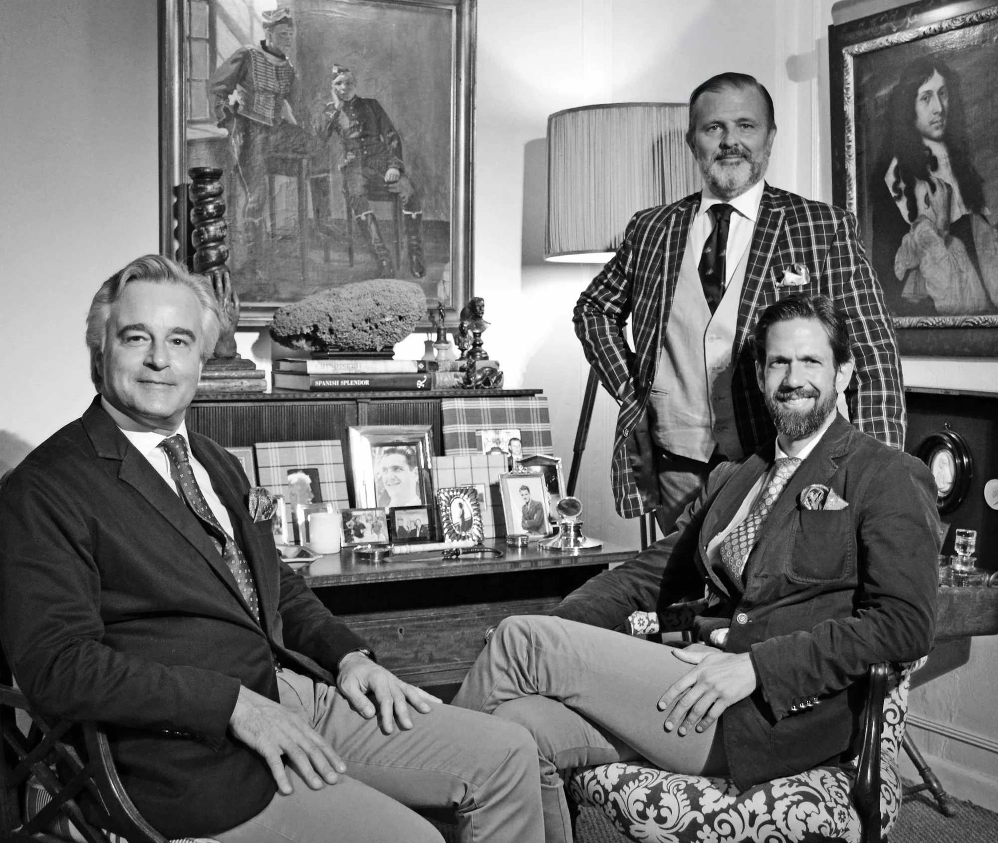 Four men in formal attire sitting and standing in a room with vintage furniture, framed photos, and artwork, in black and white.