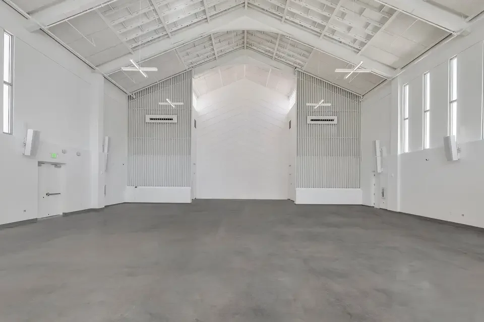 Empty white industrial or event hall with high ceiling, large windows, and minimal decor.