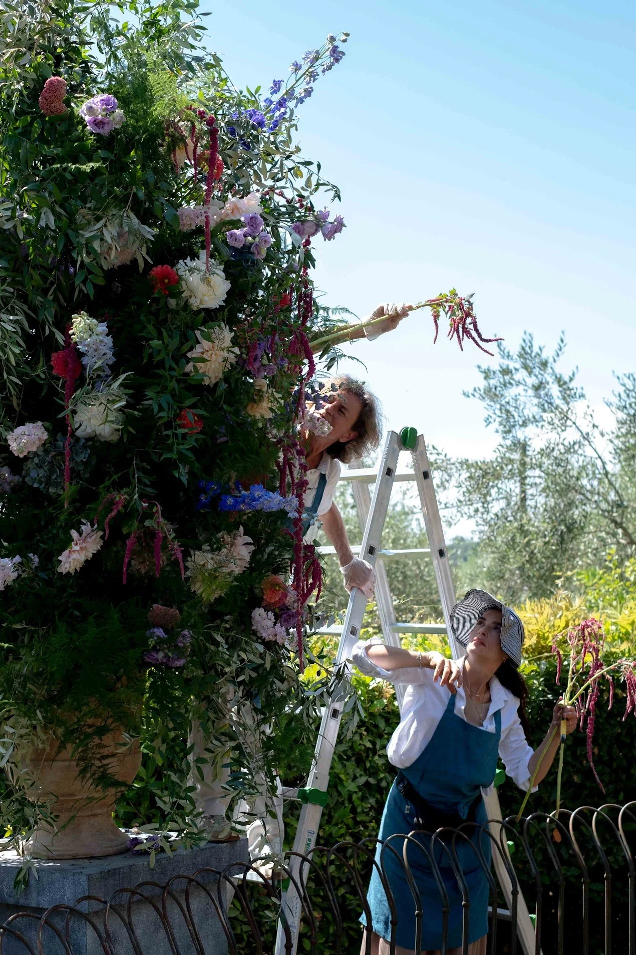 Two women tending to a large, colorful floral arrangement on a ladder outside under a clear blue sky, surrounded by greenery and a potted plant in the foreground.