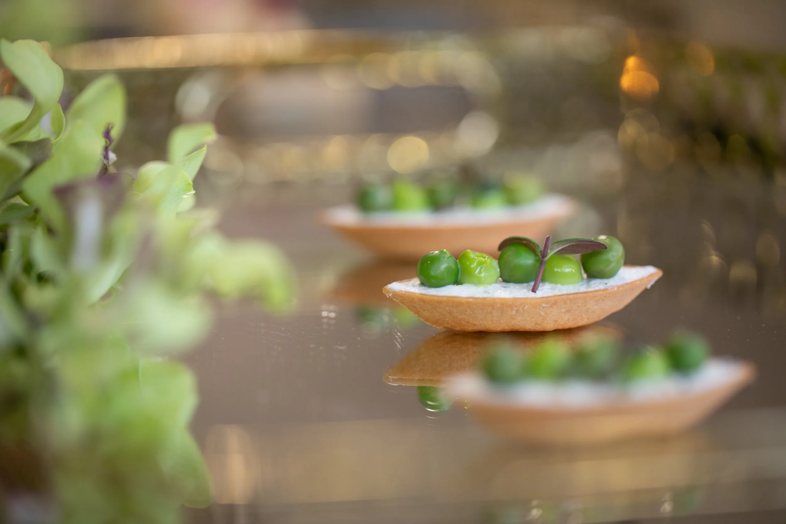 Close-up of a canapé topped with green peas and microgreens, with blurred background and reflections on a shiny surface.