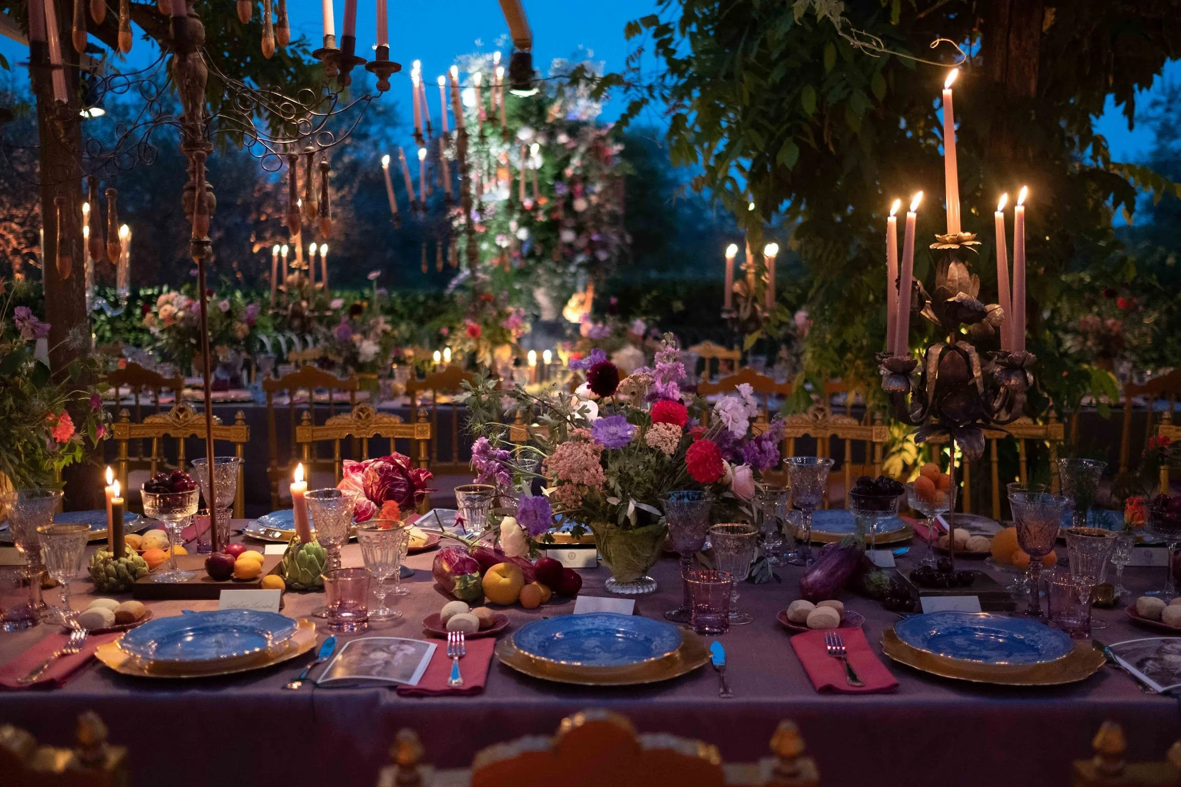 Decorated outdoor dining table set for a celebration, featuring flower arrangements, candles, and assorted desserts, with a backdrop of hanging candles and lanterns in a garden during evening.