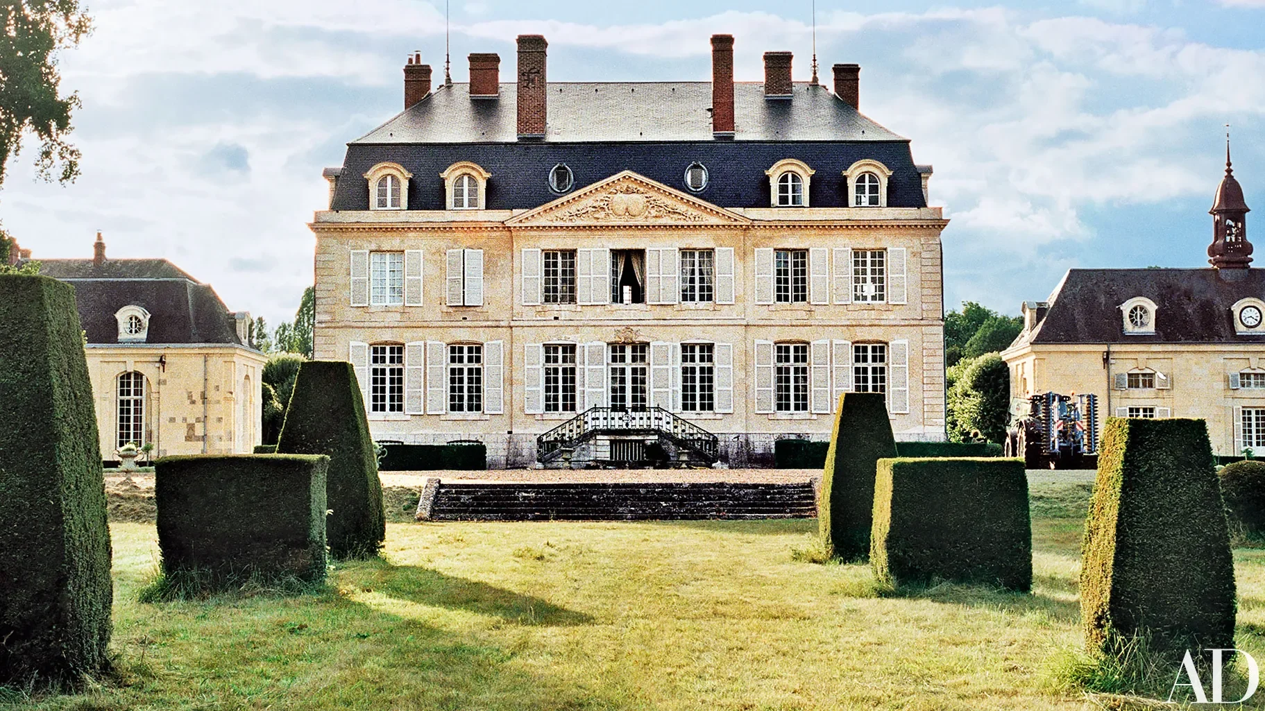 A large, elegant, three-story French chateau-style mansion with multiple chimneys, surrounded by a well-manicured lawn and trimmed hedges.
