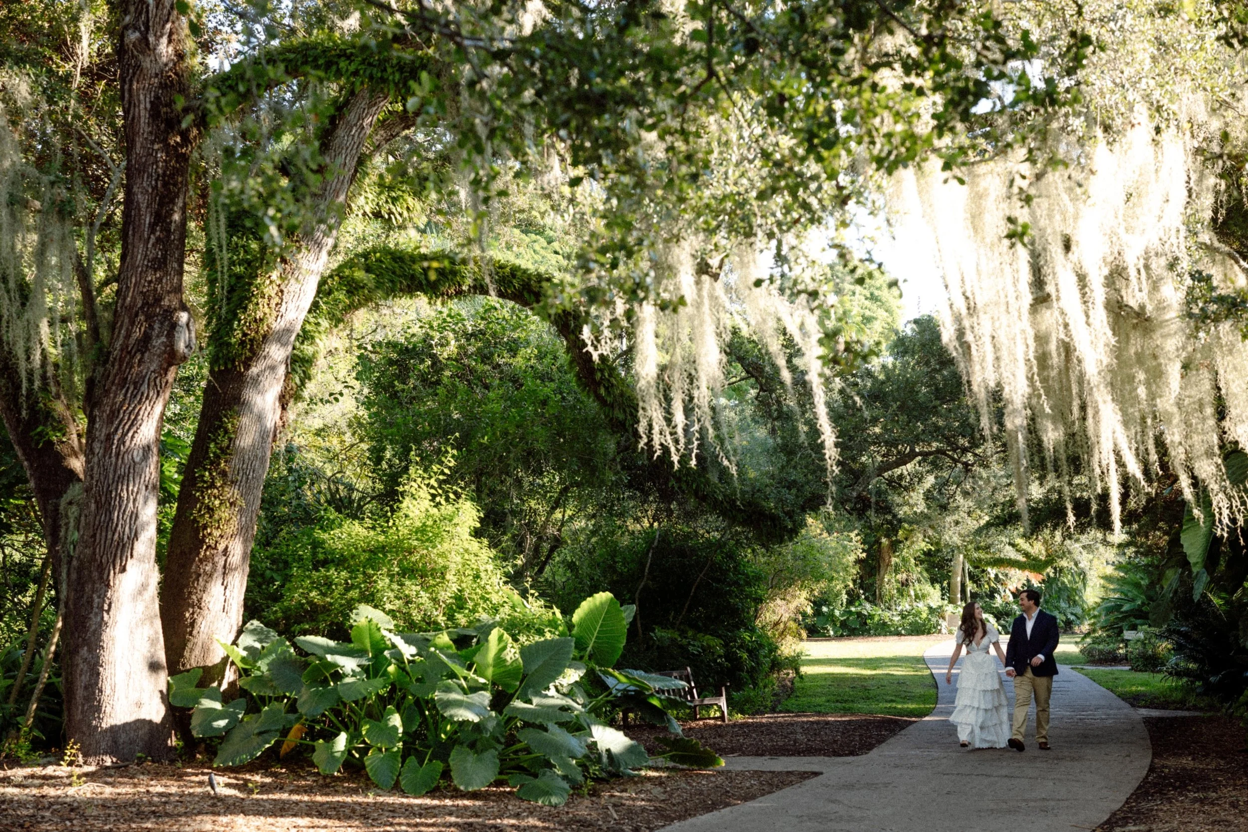 A couple dressed in wedding attire walking together in a lush, green park with trees and hanging moss, during daylight.