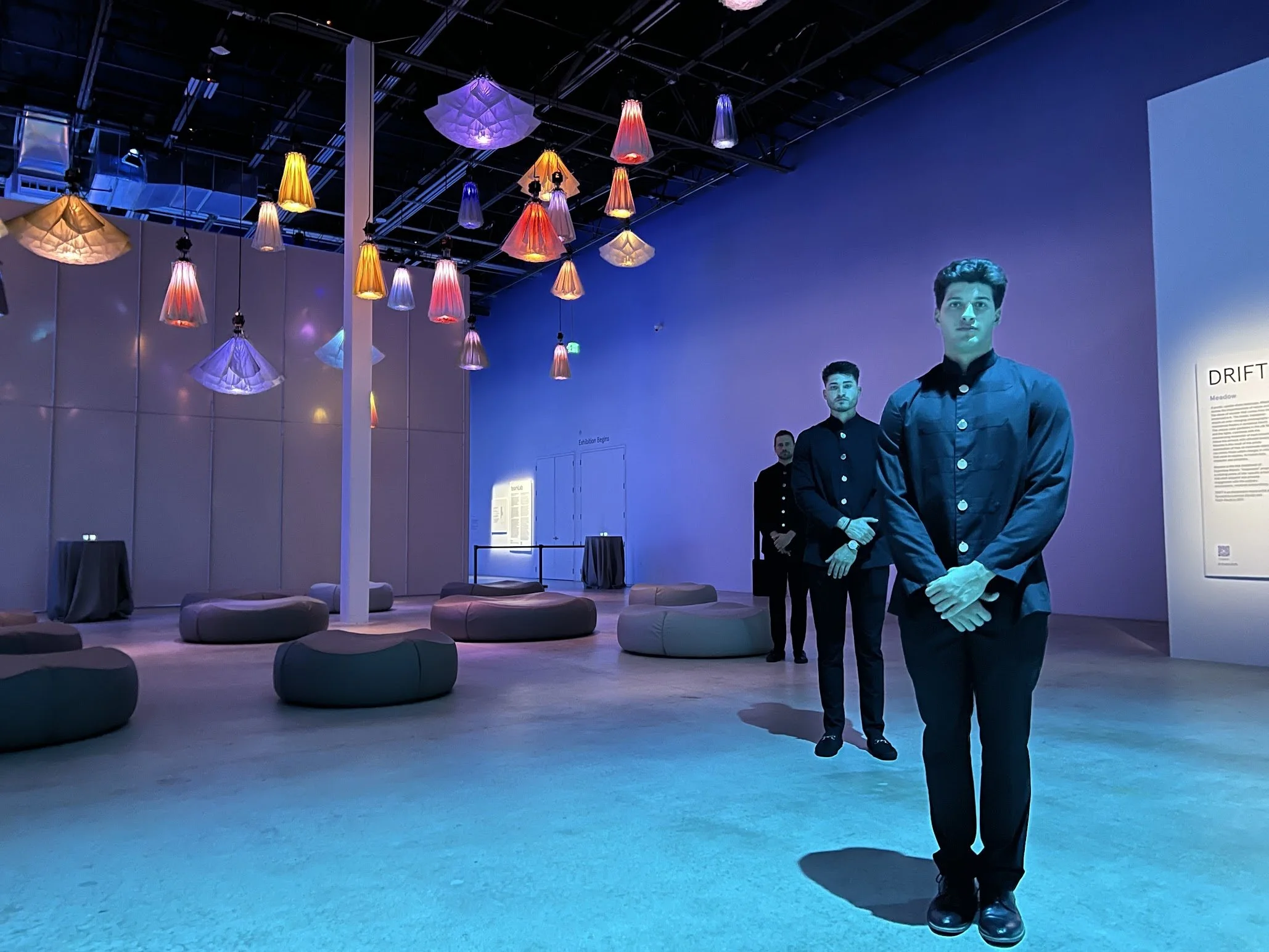 An art gallery with colorful hanging lights on the ceiling and mannequins dressed in dark clothing standing on the floor