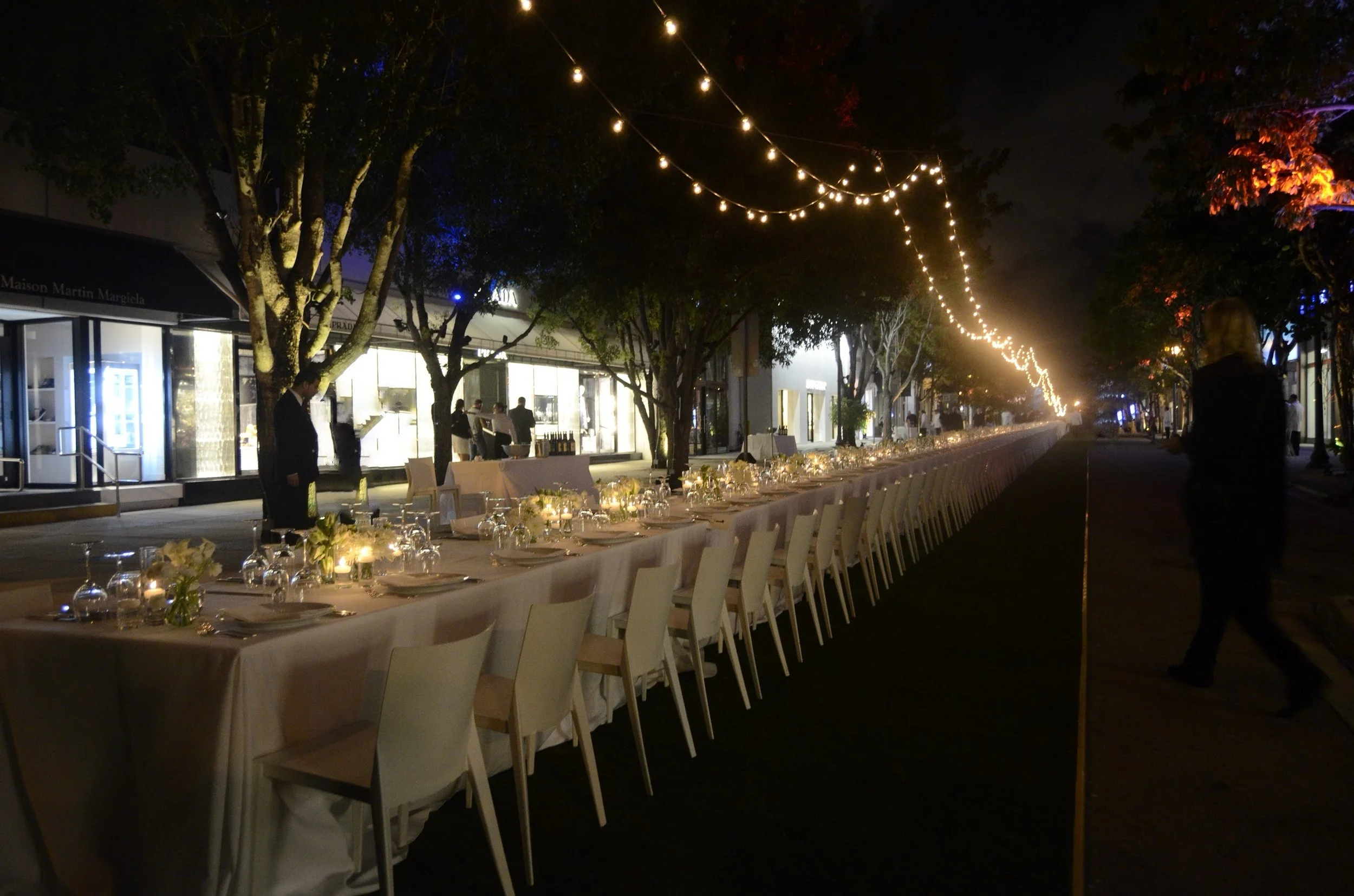 Outdoor evening dinner setup with long table covered in white cloth, decorated with flowers and candles, under string lights and trees with colorful lighting