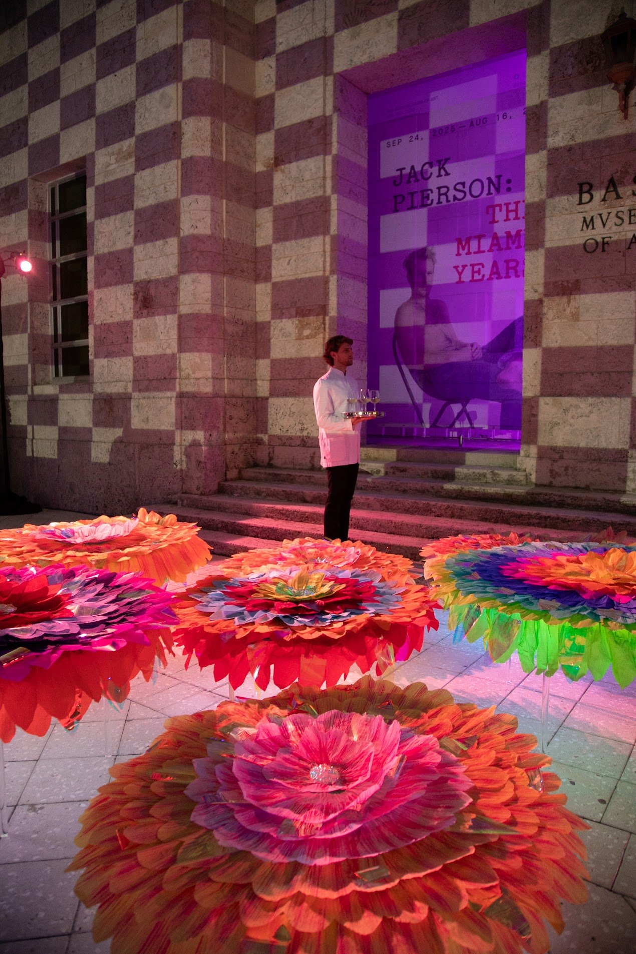 A man in a white shirt standing outdoors near a building with stone and brick pattern at night, holding a tray of glasses. The scene features large, colorful paper flower decorations arranged on the ground, and a large banner in the building's window with text about Jack Pierson and an art exhibit, illuminated with purple lighting.