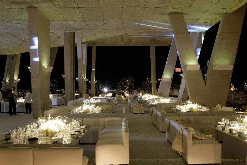 A modern outdoor dining area at night with white seating, illuminated table settings, and large concrete architecture with city lights in the background.