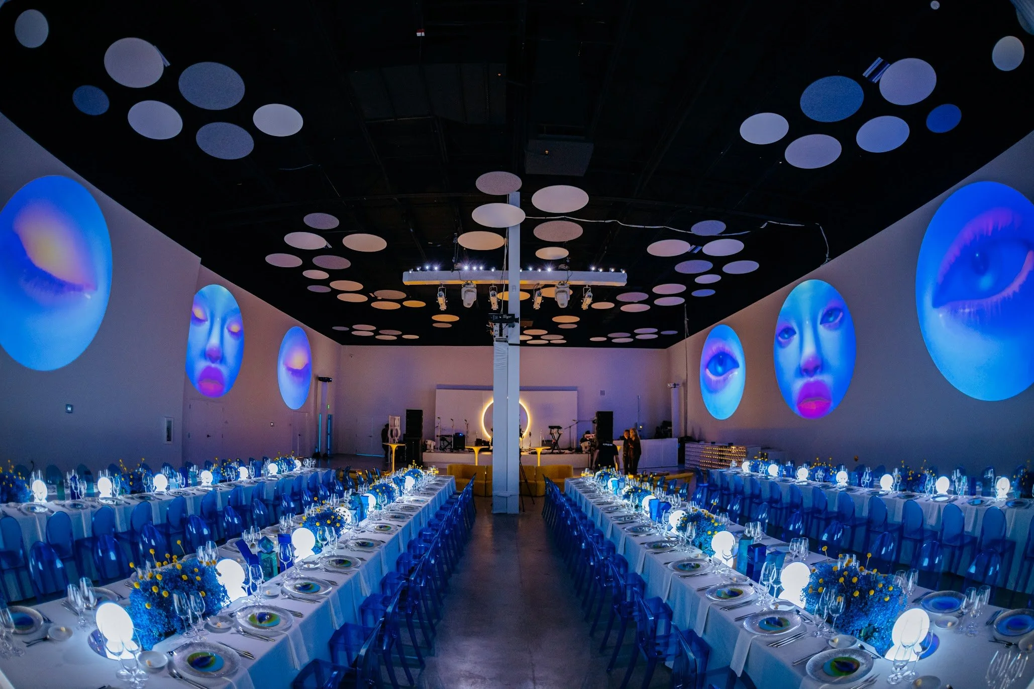 A large event hall decorated for a formal occasion with long dining tables set with white tablecloths, blue chairs, and centerpieces of yellow and blue flowers. The ceiling has circular light fixtures and projected images of blue faces with closed eyes on the walls. Stage equipment is visible at the far end.