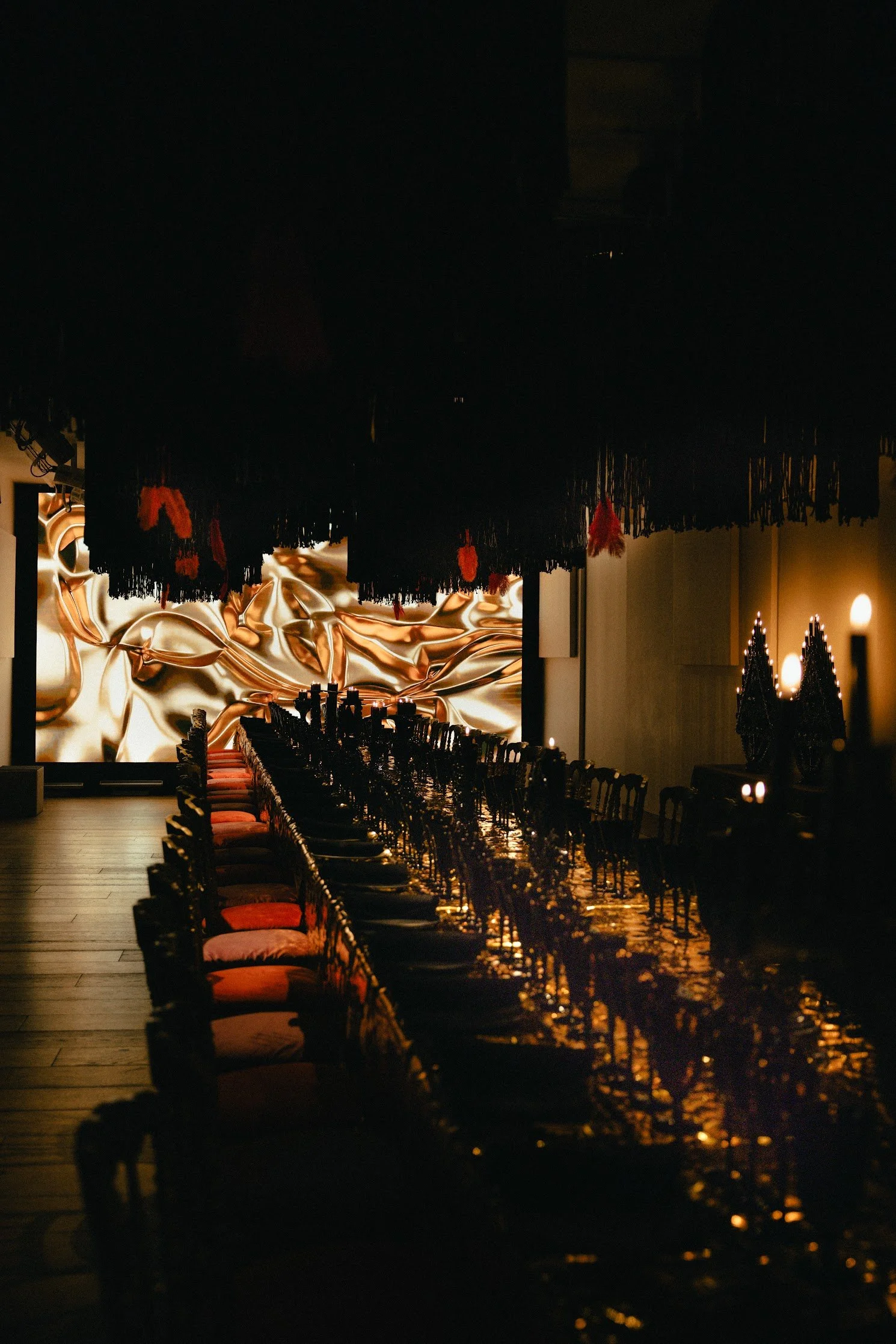 Empty long dining table with red and black chairs in a dimly lit restaurant with large abstract gold wall art and hanging lamps.