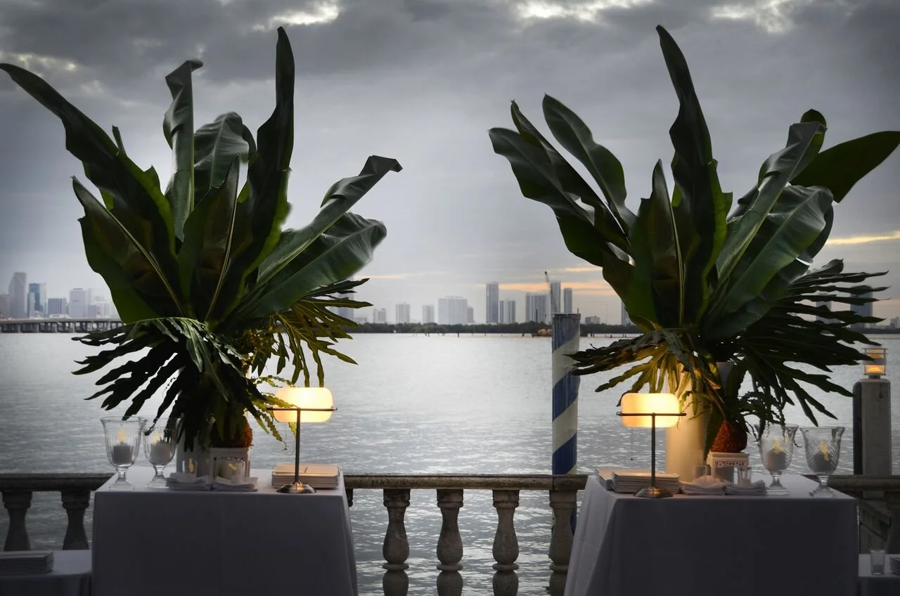 Two tables set with tableware and lamps, decorated with large tropical plants, overlooking a body of water with a city skyline in the background at dusk.