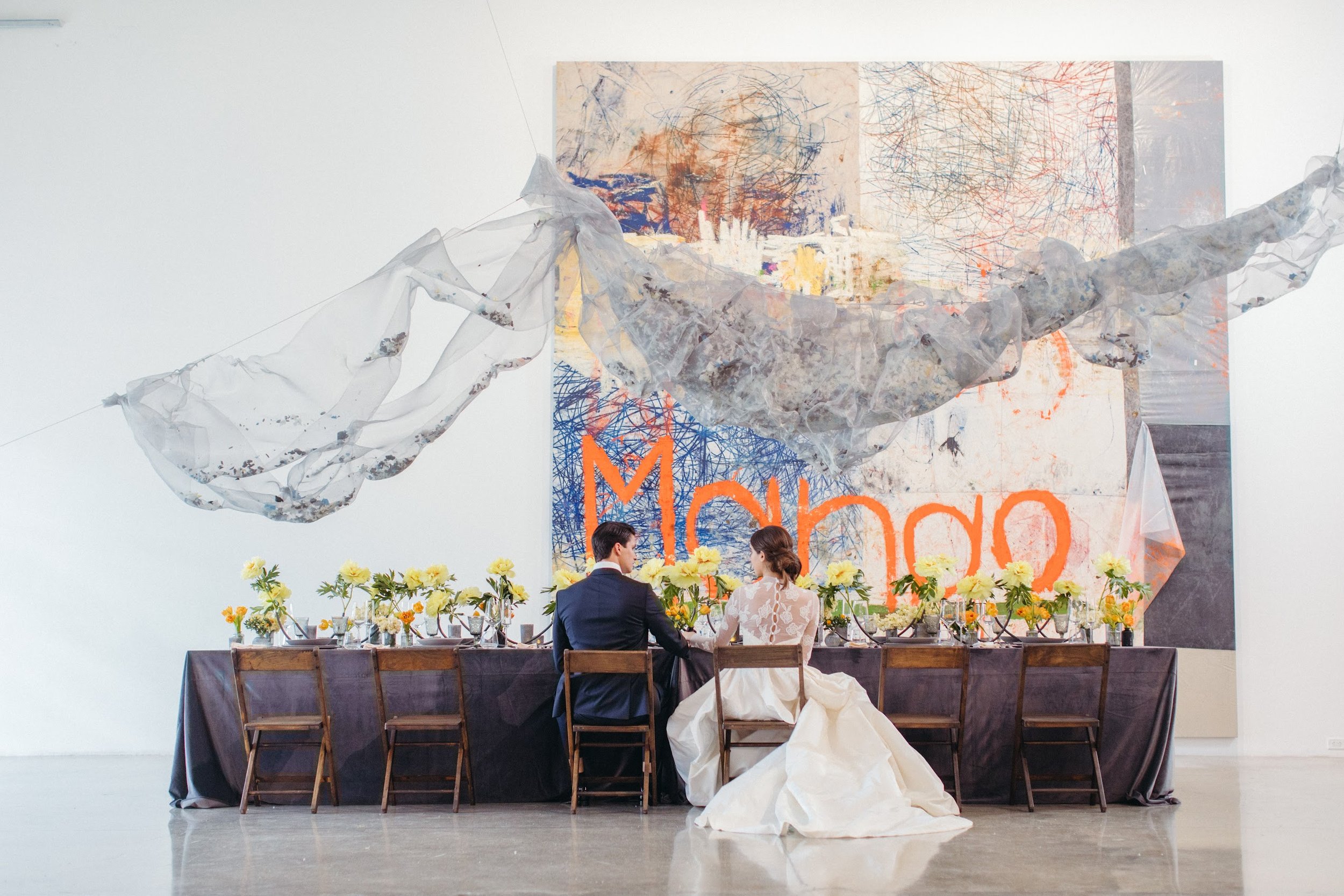A wedding reception setup with a long table decorated with yellow flowers, where a bride and groom are seated facing each other, holding hands. Behind them, a large abstract artwork with colorful scribbles is displayed on the wall.