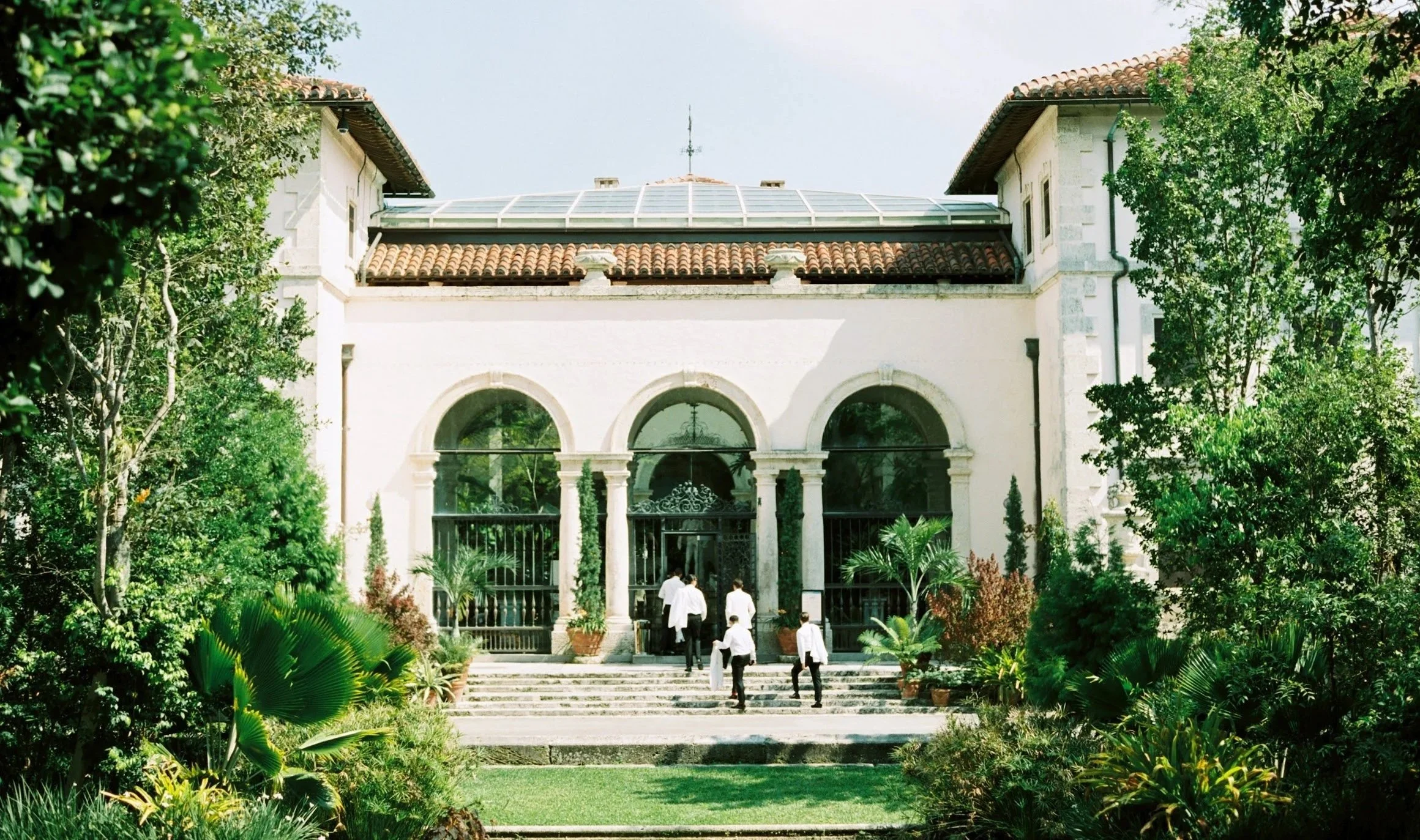 A historic mansion with arched windows and a tile roof, surrounded by lush greenery and a group of people walking toward the entrance.