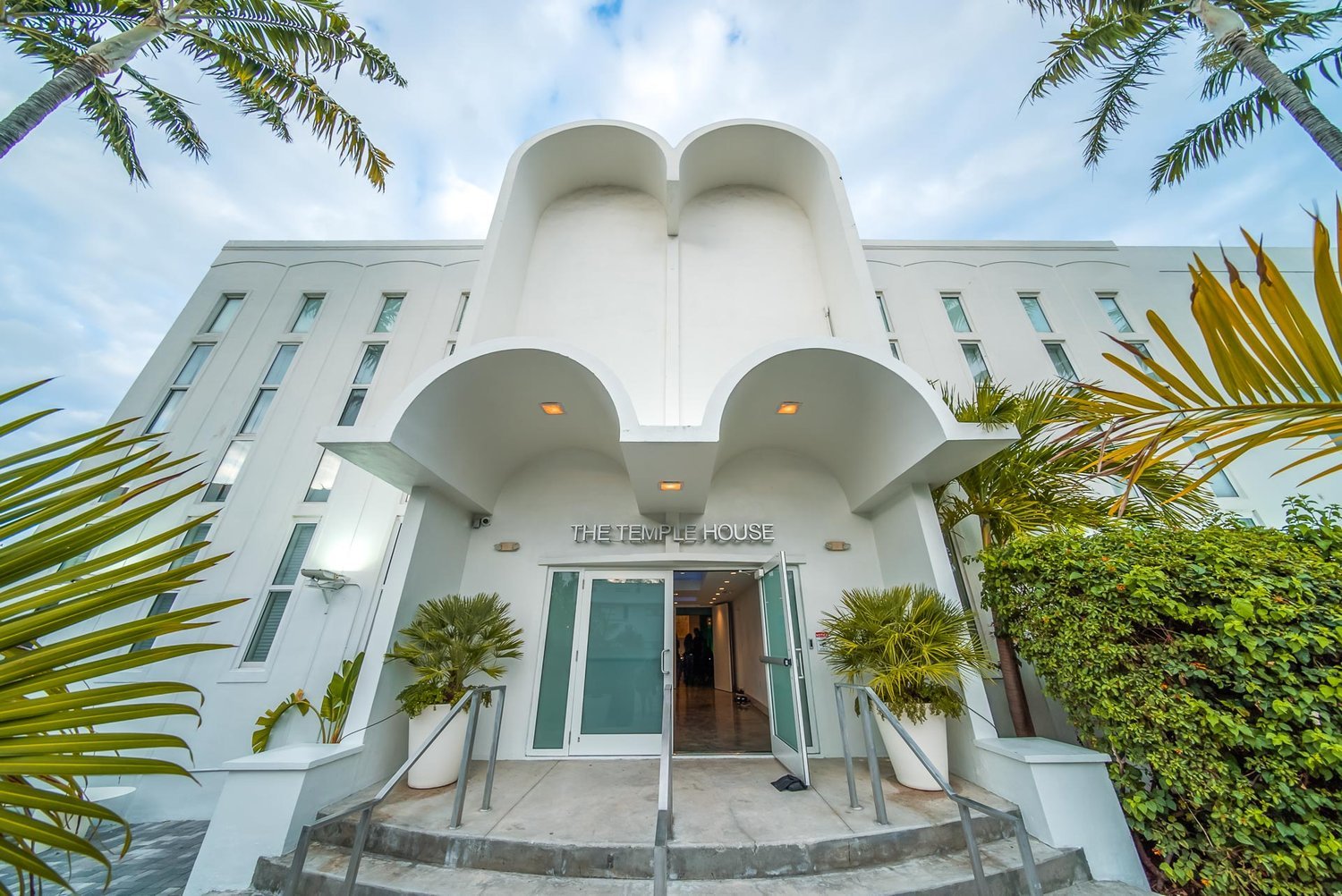 Exterior view of The Temple House, a white building with an architectural design featuring rounded arches and tall narrow windows, surrounded by palm trees and greenery, with a blue sky overhead.