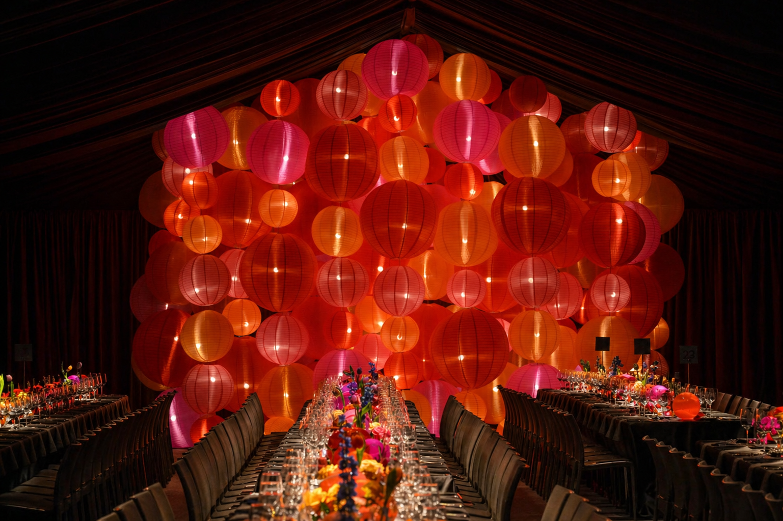 A banquet hall decorated with a large cluster of pink, red, and orange paper lanterns hanging from the ceiling. The tables are set with glasses, dinnerware, and colorful flower centerpieces.
