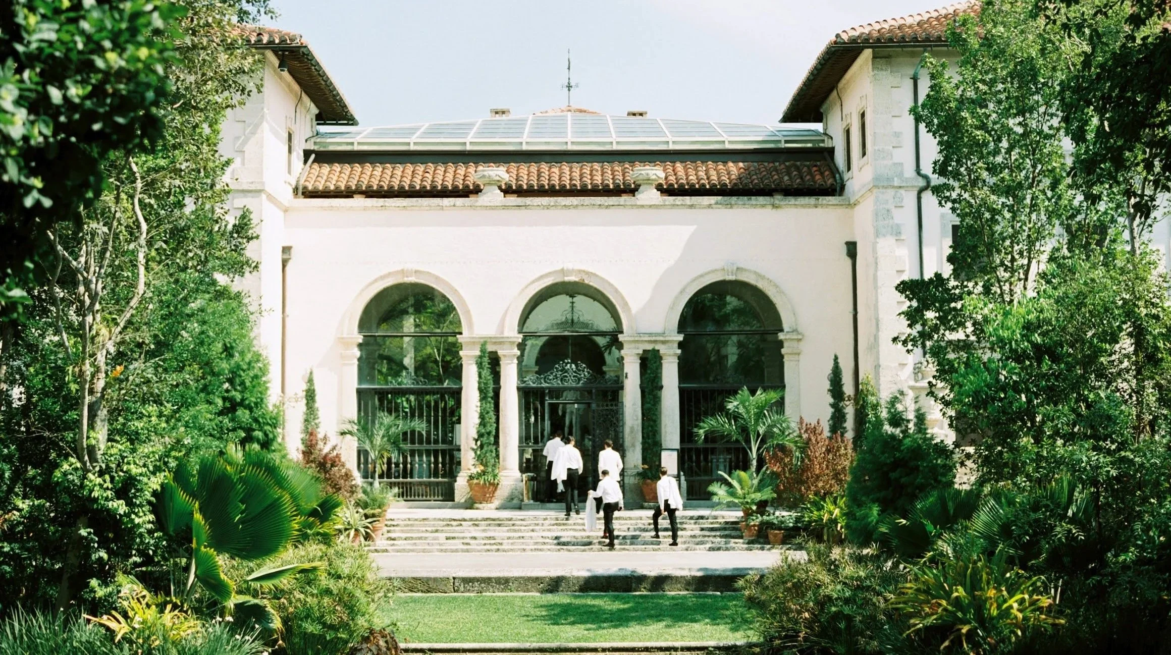 A building with white walls, arched windows, a tiled roof, and surrounded by lush greenery, with people walking toward the entrance.